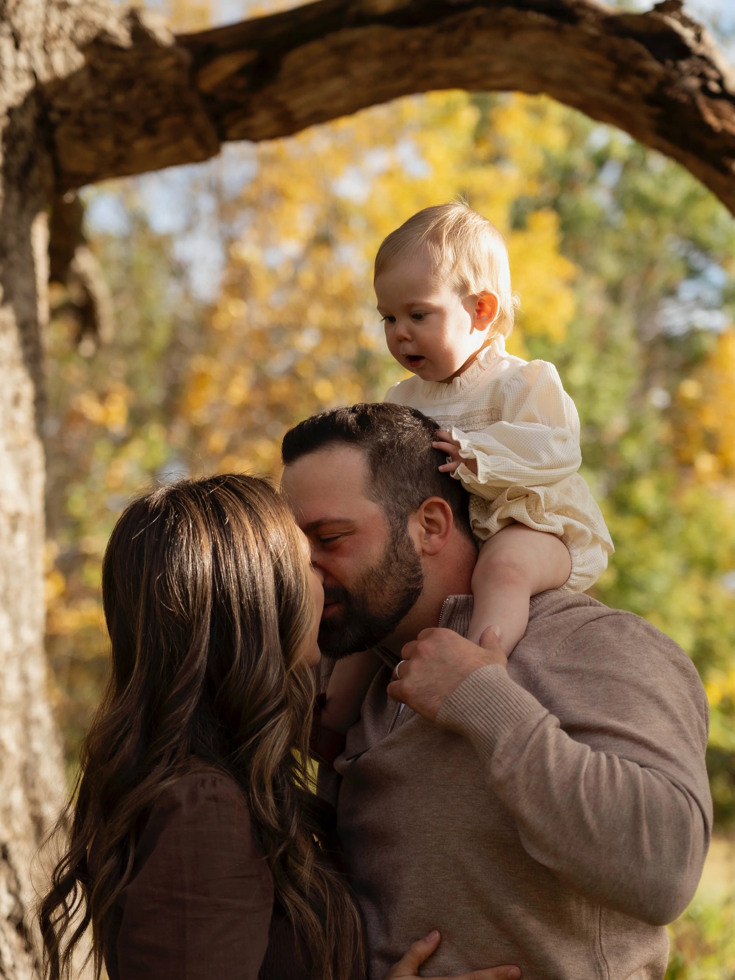 A family of three, a man, a woman, and a young girl, sharing a kiss outdoors during fall, with colorful autumn leaves in the background.