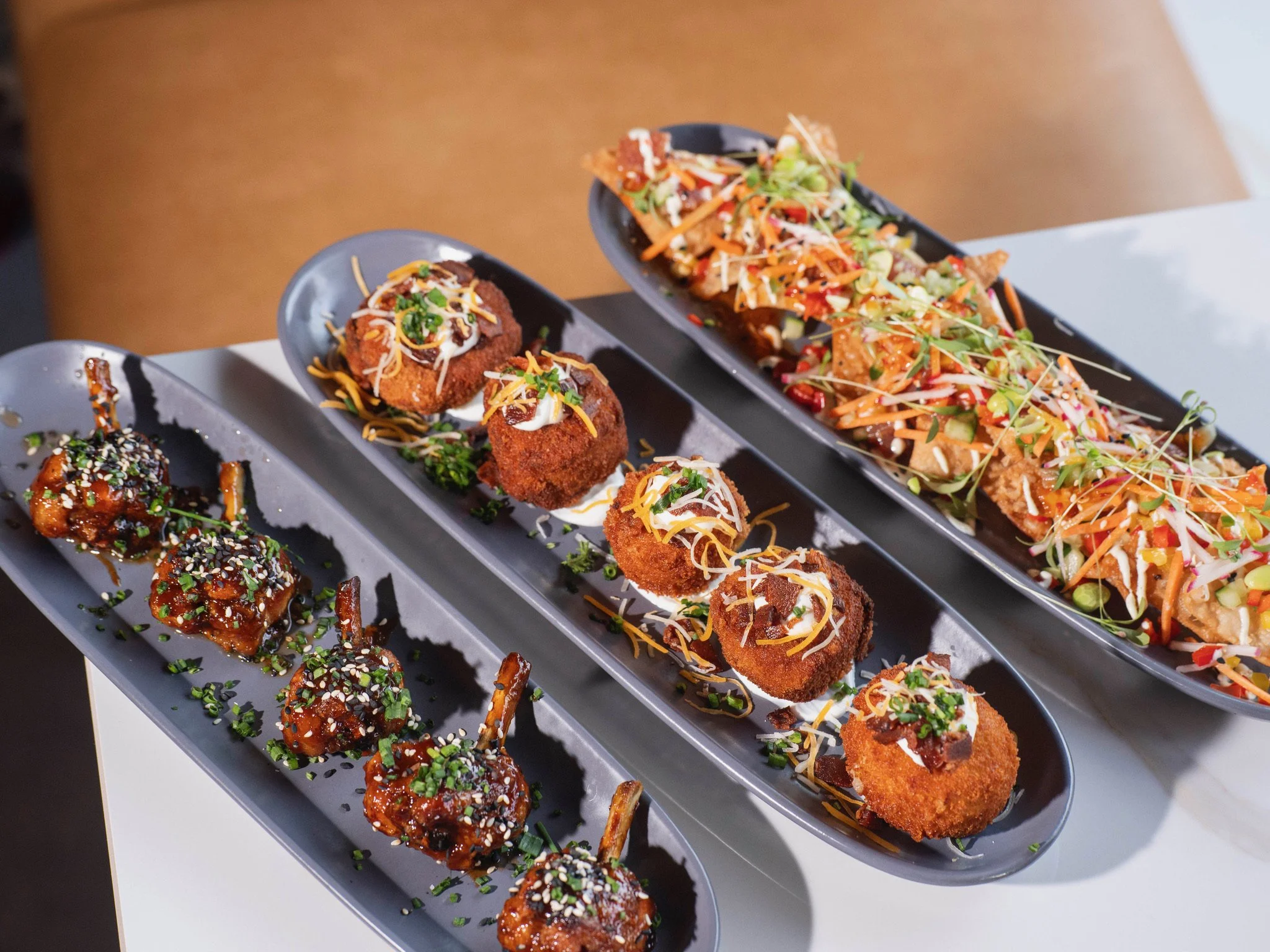 Three black elongated plates with Asian-inspired appetizers: the left plate has glazed chicken drumettes garnished with sesame seeds and chopped green onions; the middle plate has four croquettes topped with sauces, shredded cheese, and chopped herbs