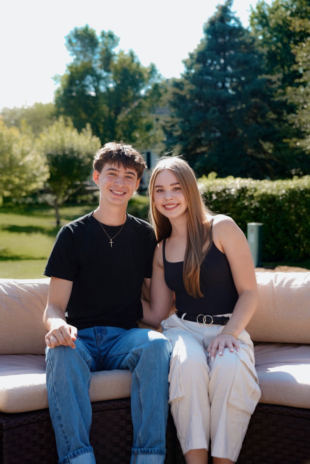 Two young people, a male and a female, sitting on a beige sofa outdoors, smiling at the camera with trees and a sunny sky in the background.