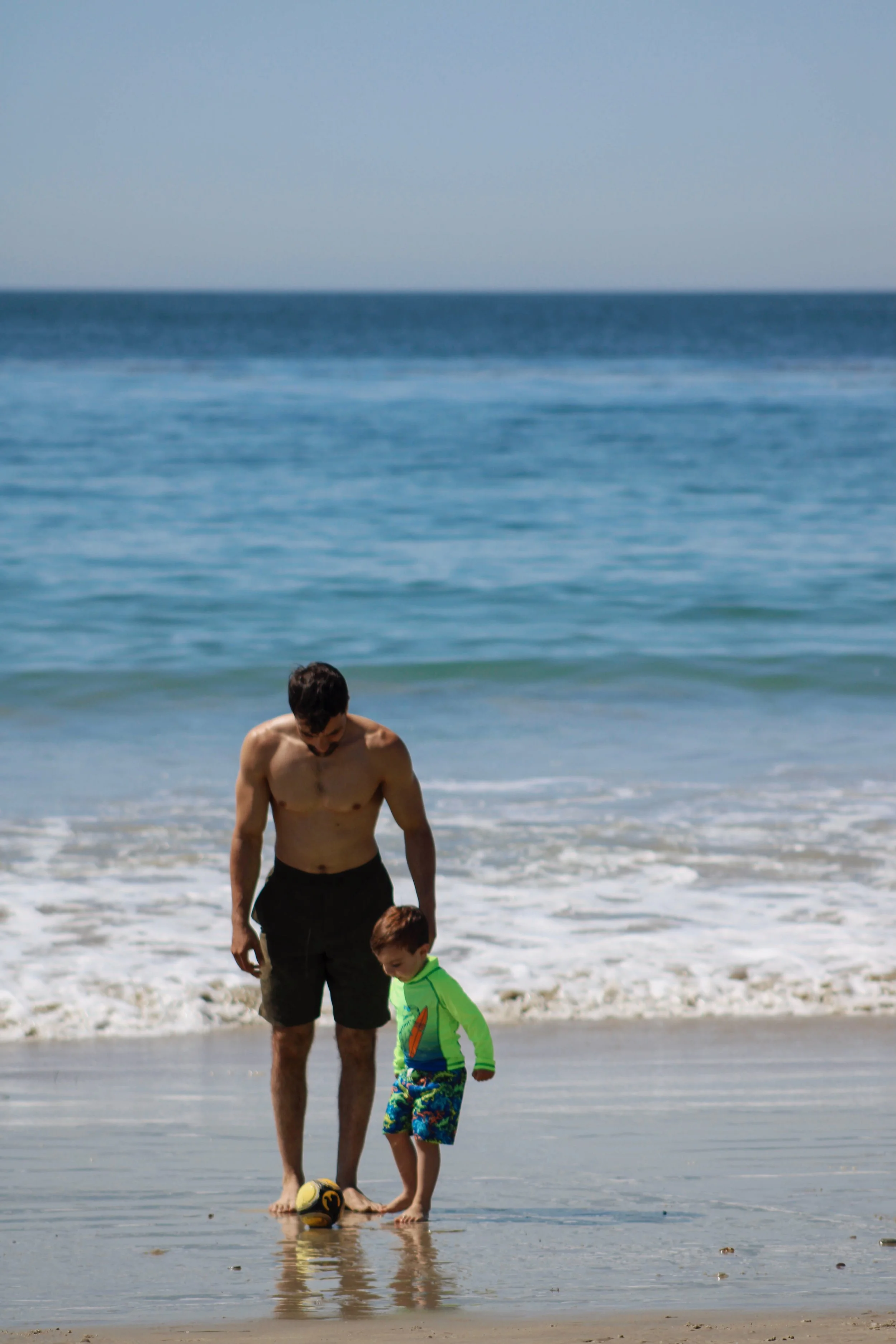 A man and a young boy playing soccer on the beach near the water's edge.