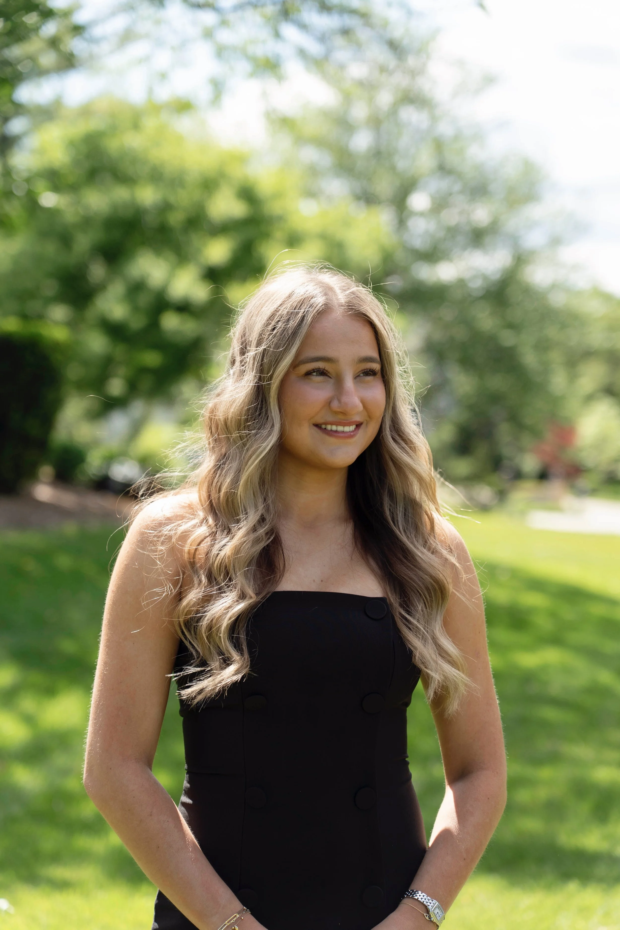 Girl with long wavy blonde hair wearing a black sleeveless dress standing outdoors on a sunny day with green trees and grass in the background.