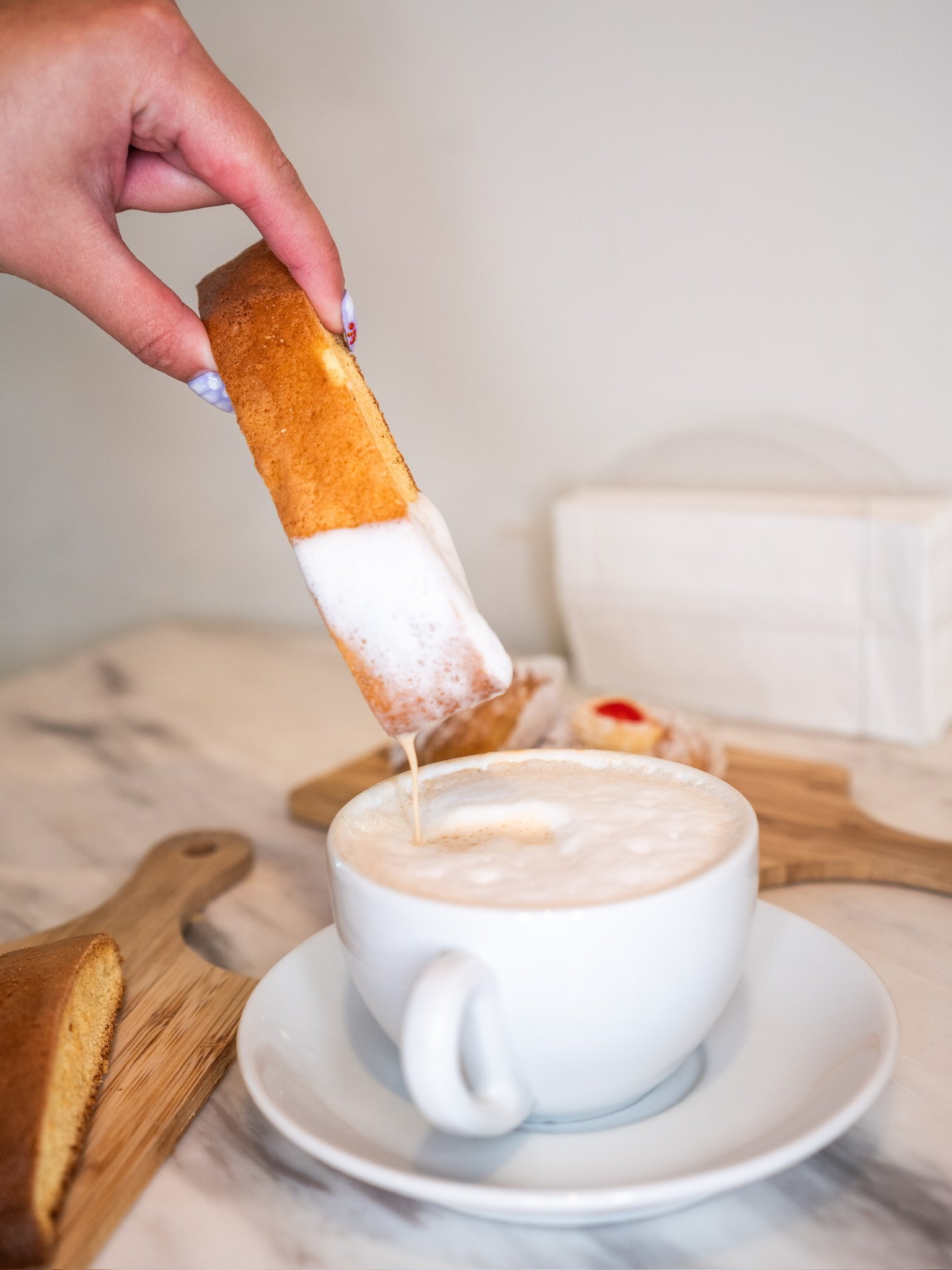 Person dipping a toasted bread stick into a cup of frothy coffee or hot beverage on a white saucer, with a wooden board and more bread sticks in the background.