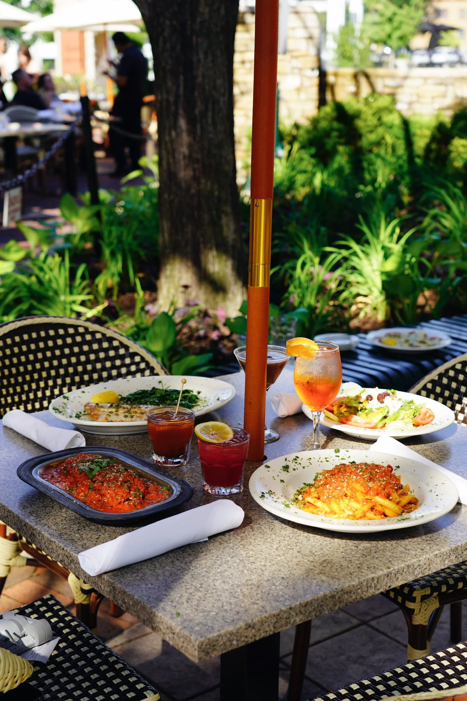 Outdoor restaurant table with pasta, salads, and drinks, tables, chairs, greenery, and people in the background.