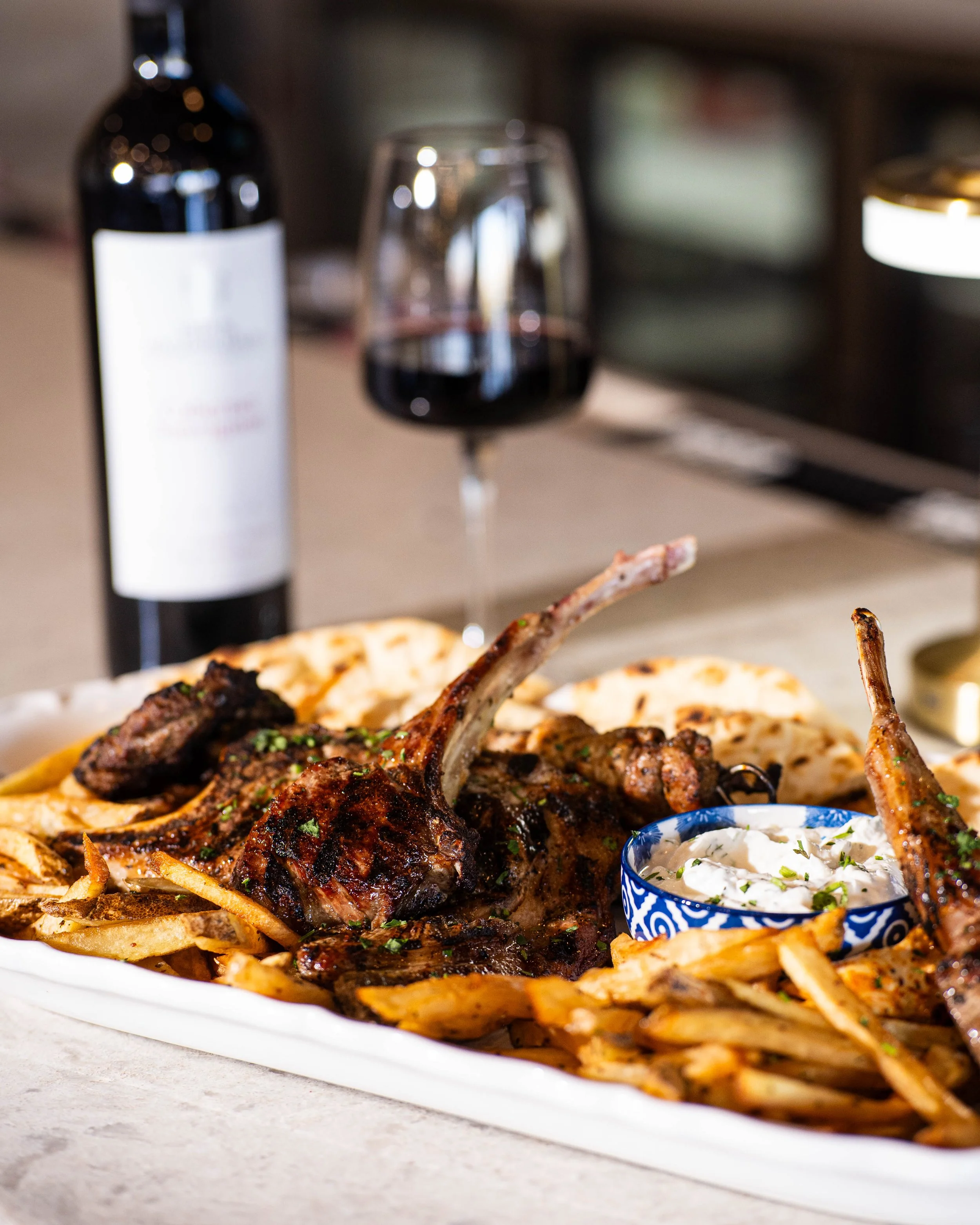 A plate of grilled lamb chops served with French fries, pita bread, a small bowl of creamy dipping sauce, and a glass of red wine with a bottle in the background.