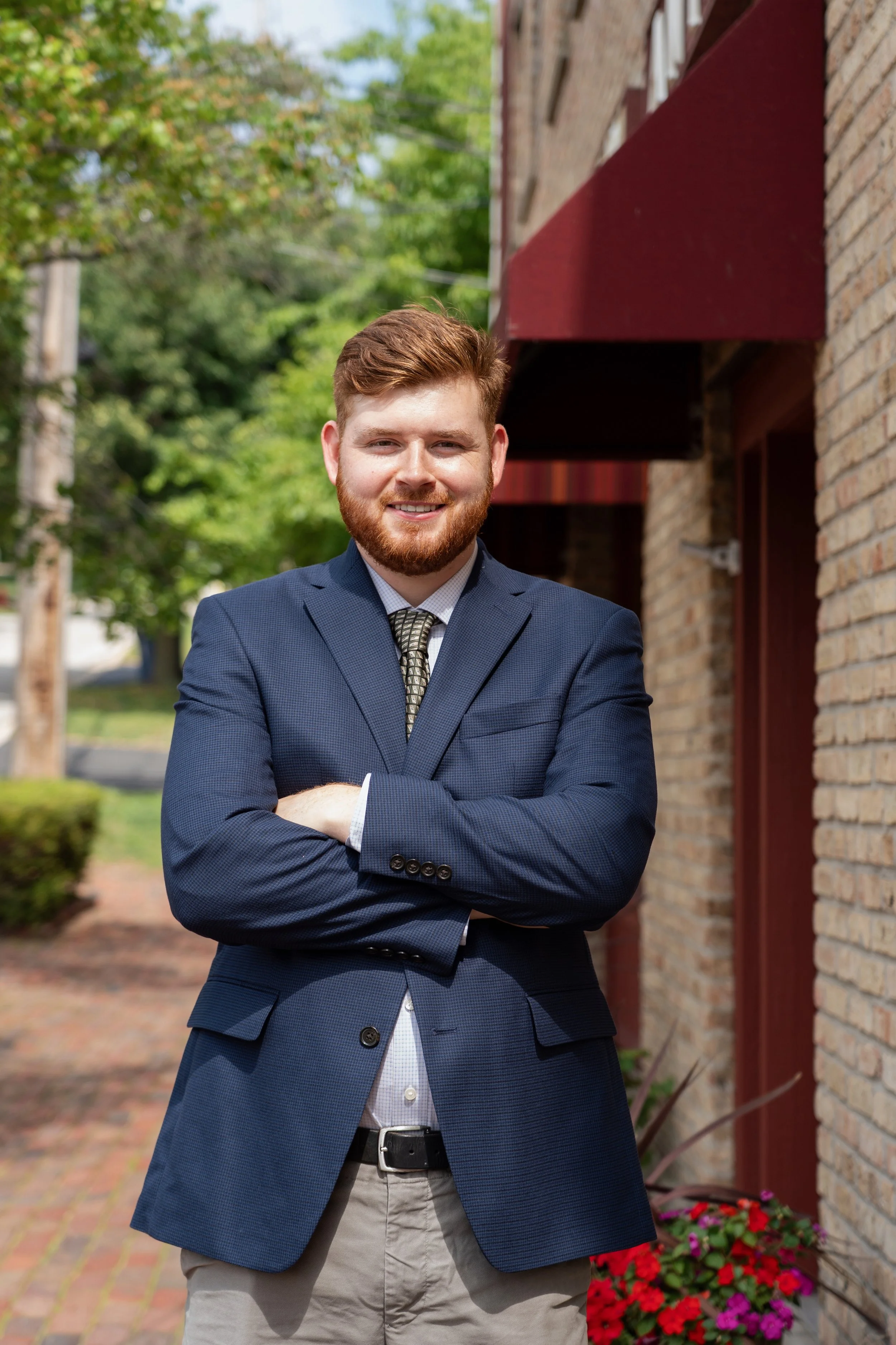 A young man with red hair and a beard is smiling, standing outdoors with crossed arms, wearing a blue suit jacket, a white shirt, and a patterned tie, in front of a brick building with green trees in the background.