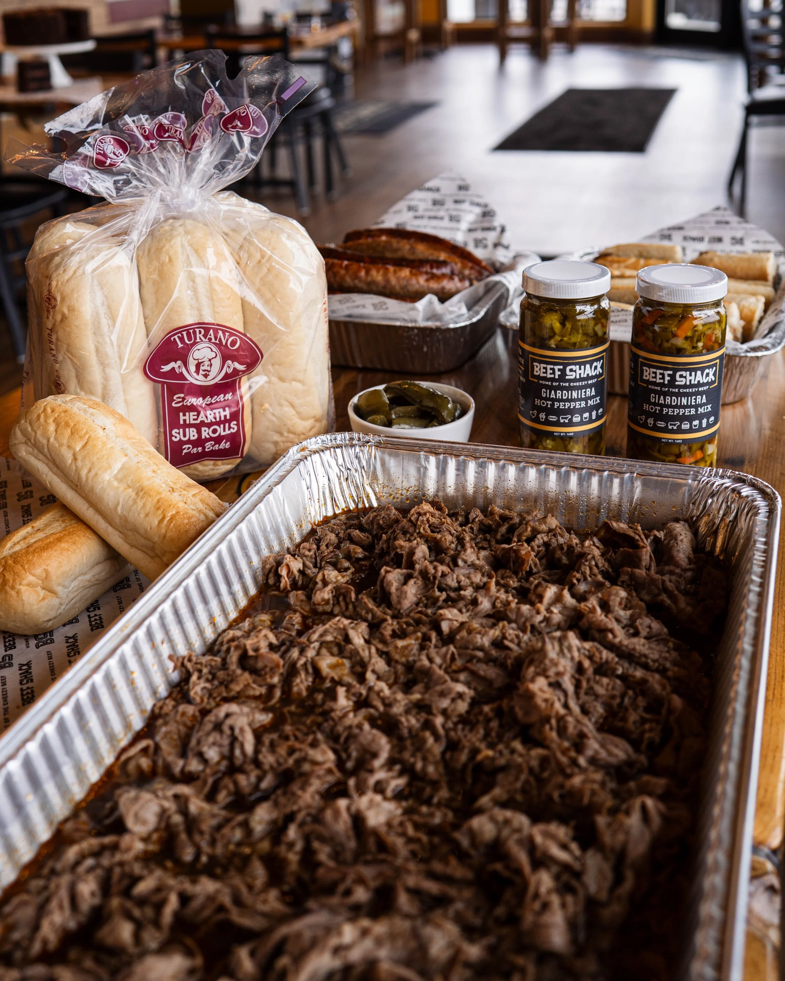 A table filled with cooked shredded beef, sub rolls, hot dogs, pickles, and jars of hot pepper mix, set in a cozy restaurant or dining area.