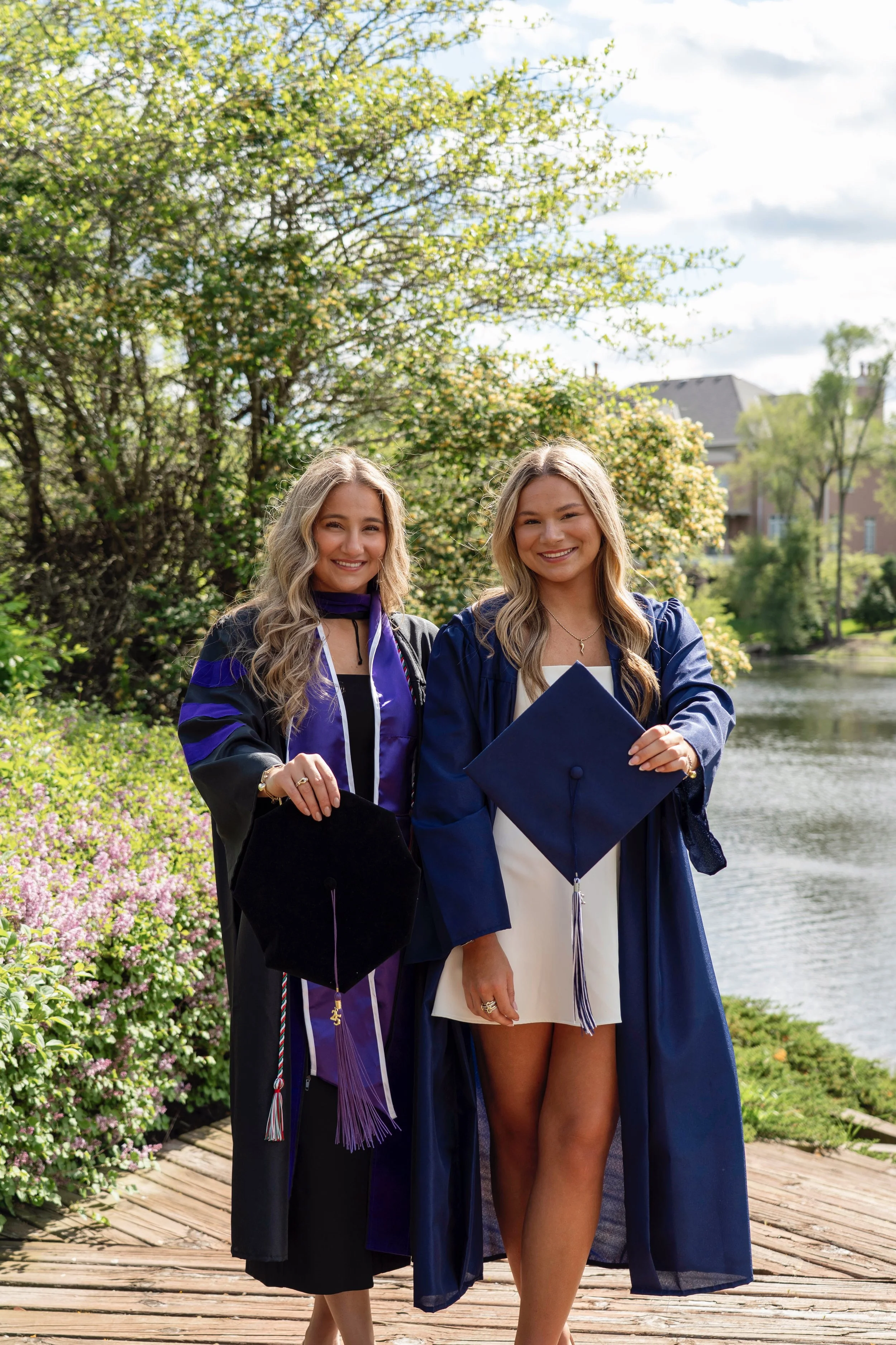 Two young women in graduation caps and gowns smiling outdoors near a water body with trees and shrubs in the background.