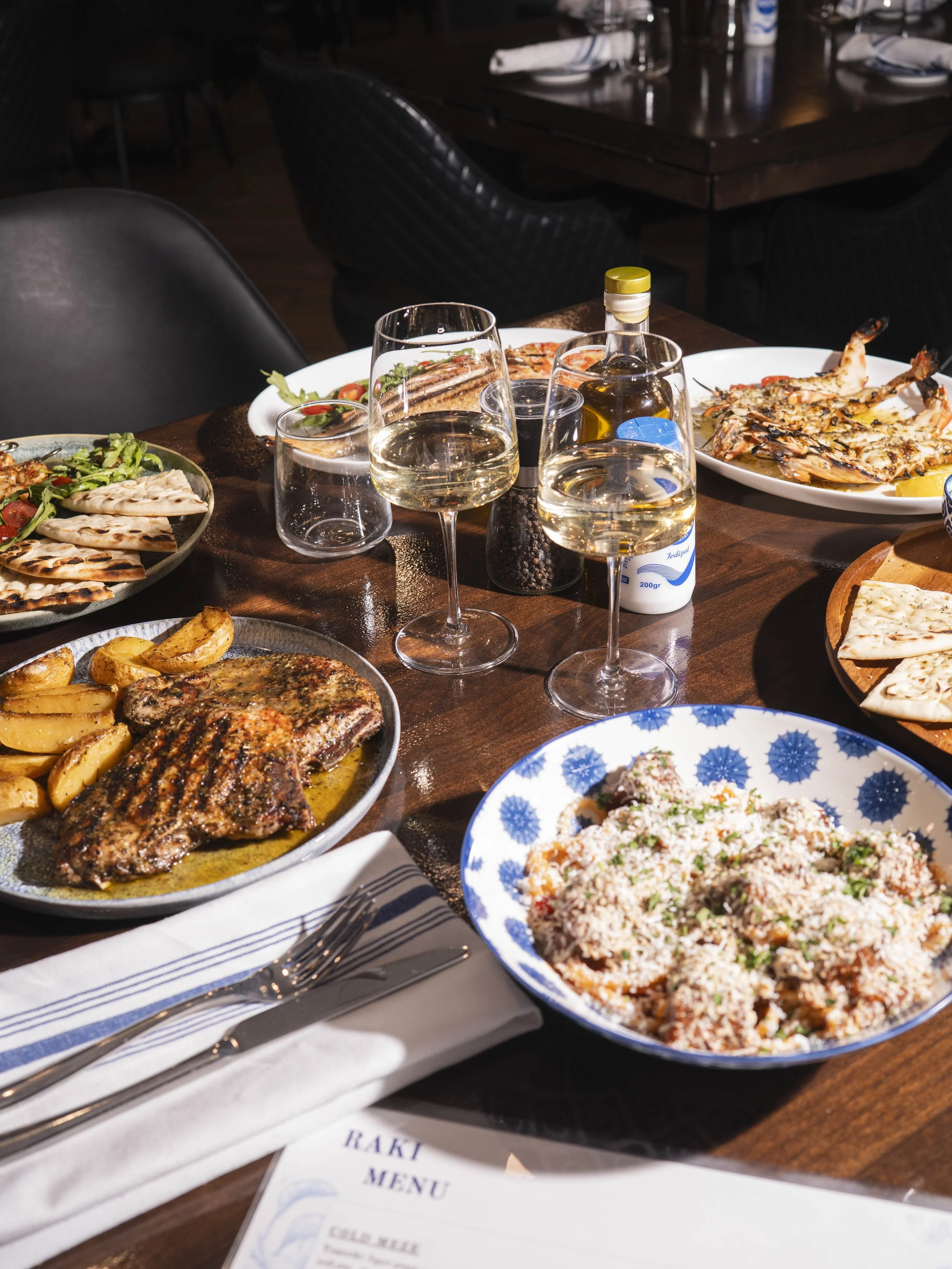 Dinner table with plates of grilled meats, roasted potatoes, salad, rice dish, pink and white wine, water, and condiments.