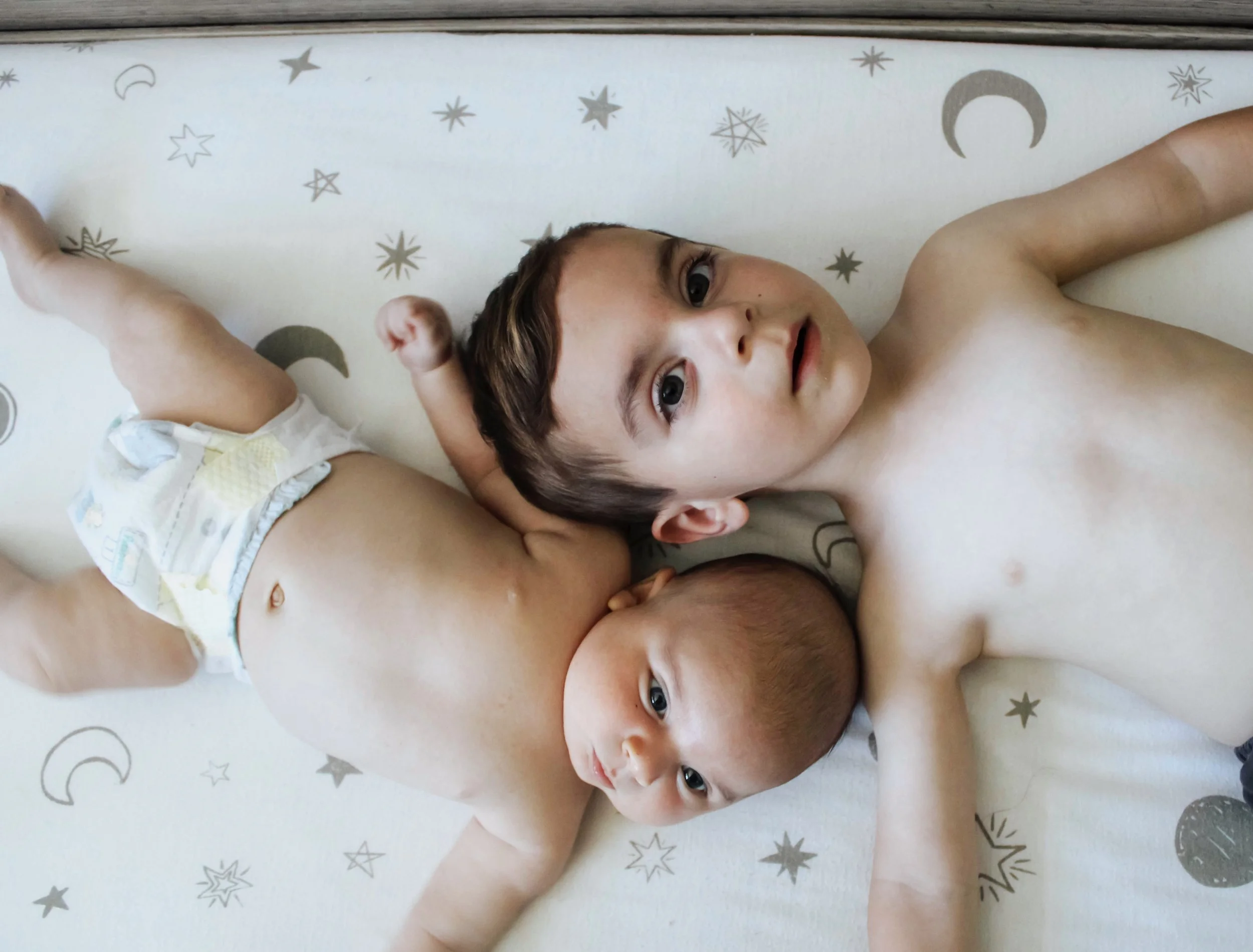 Two young children, one with dark hair and one with lighter hair, lying down on a mattress with a celestial pattern, looking up at the camera.