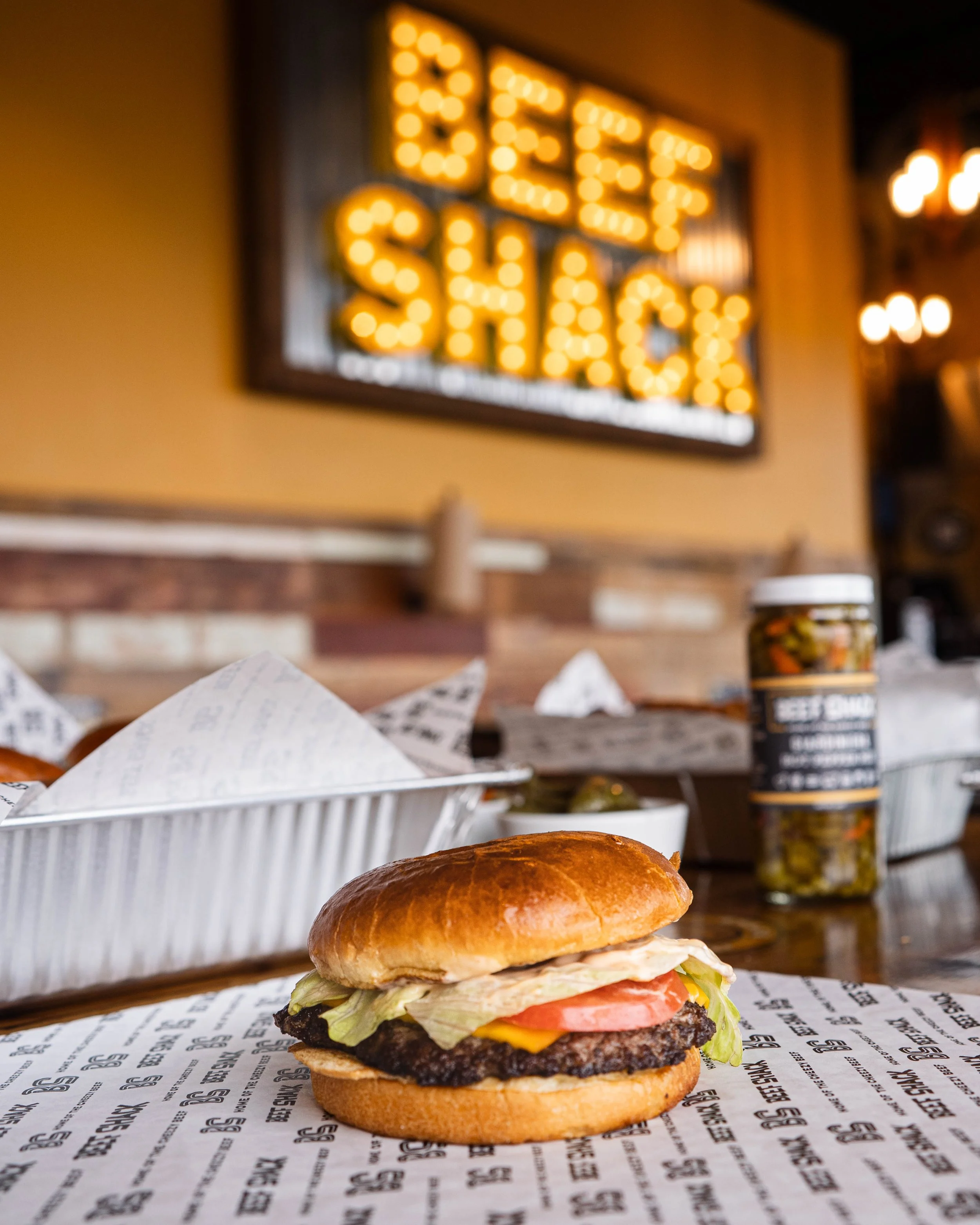 Close-up of a cheeseburger with lettuce, tomato, pickles, and cheese on a bun, on a paper-lined tray, with a bottle of pickles and a basket with paper on a wooden table, in a restaurant with a bright yellow "Beer Shack" sign in the background.