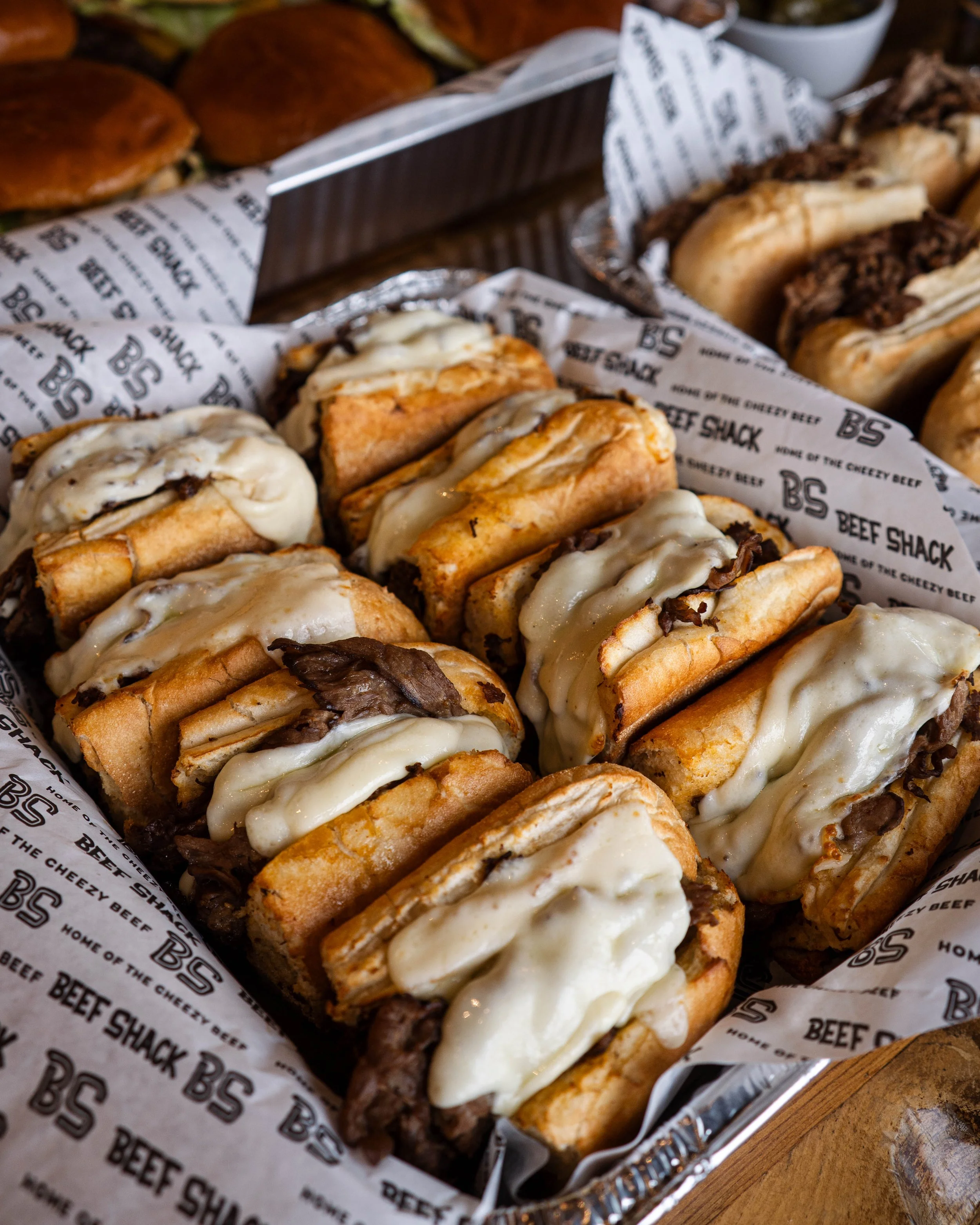 A tray of cheeseburgers with melted cheese and beef patties on sandwich buns, placed on branded paper in a metal serving tray.