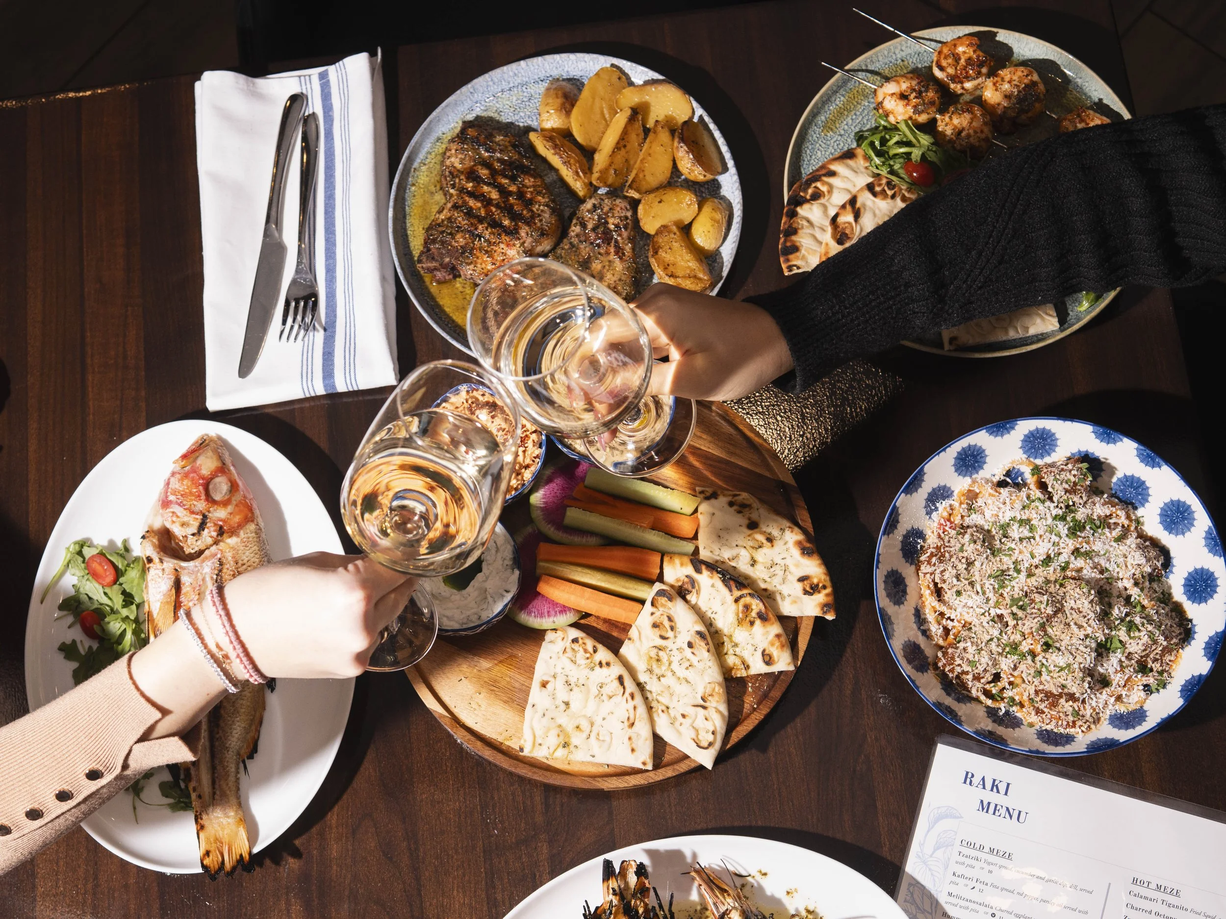 Diverse dishes of Middle Eastern and Mediterranean cuisine on a dark wooden table, including grilled meats, vegetables, flatbreads, a fish, a salad with rice, and glasses of white wine being clinked in a toast.