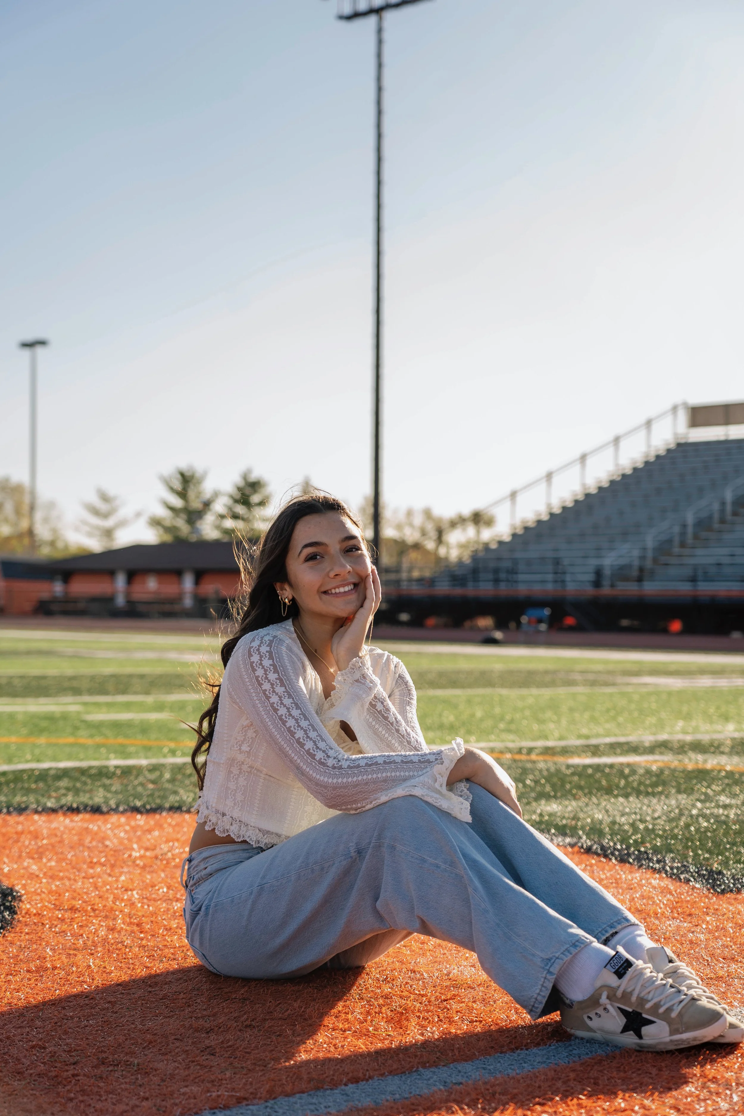 Young woman sitting on a running track, smiling at the camera with her hand on her face, wearing a white long sleeve top, light blue jeans, and sneakers, with a sports field and bleachers in the background during sunlight.