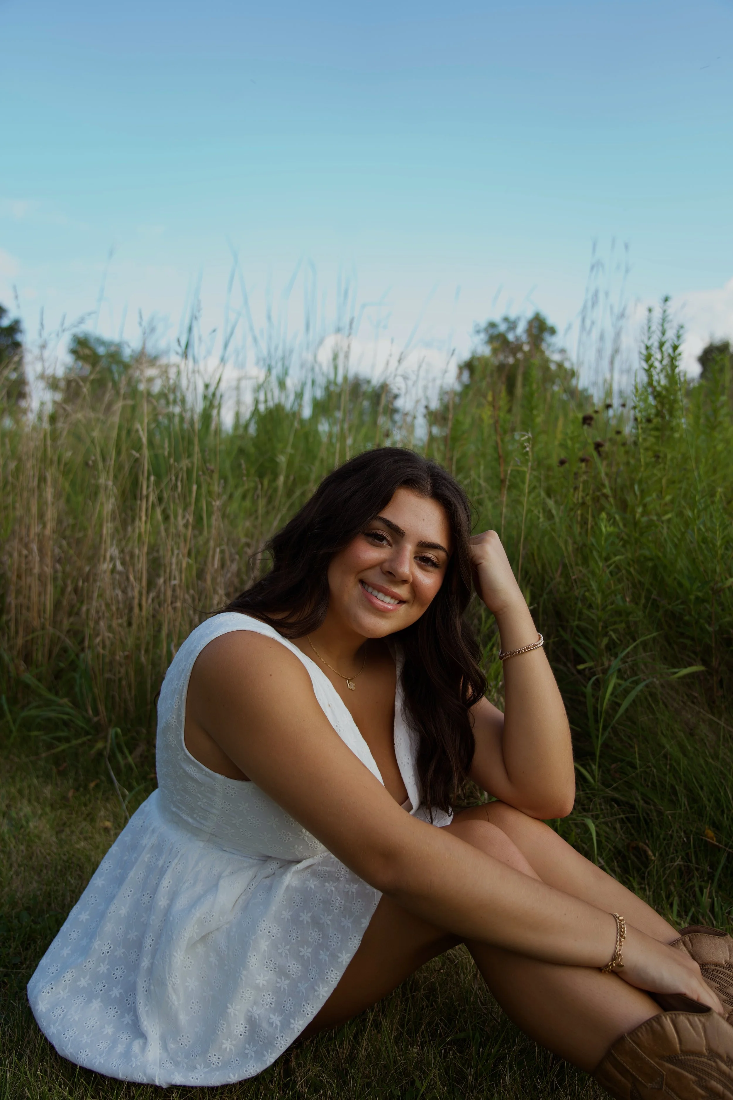 A young woman with long dark hair sitting on grass in a white dress, smiling with her hand resting on her head, surrounded by tall green grass and a blue sky with some clouds in the background.