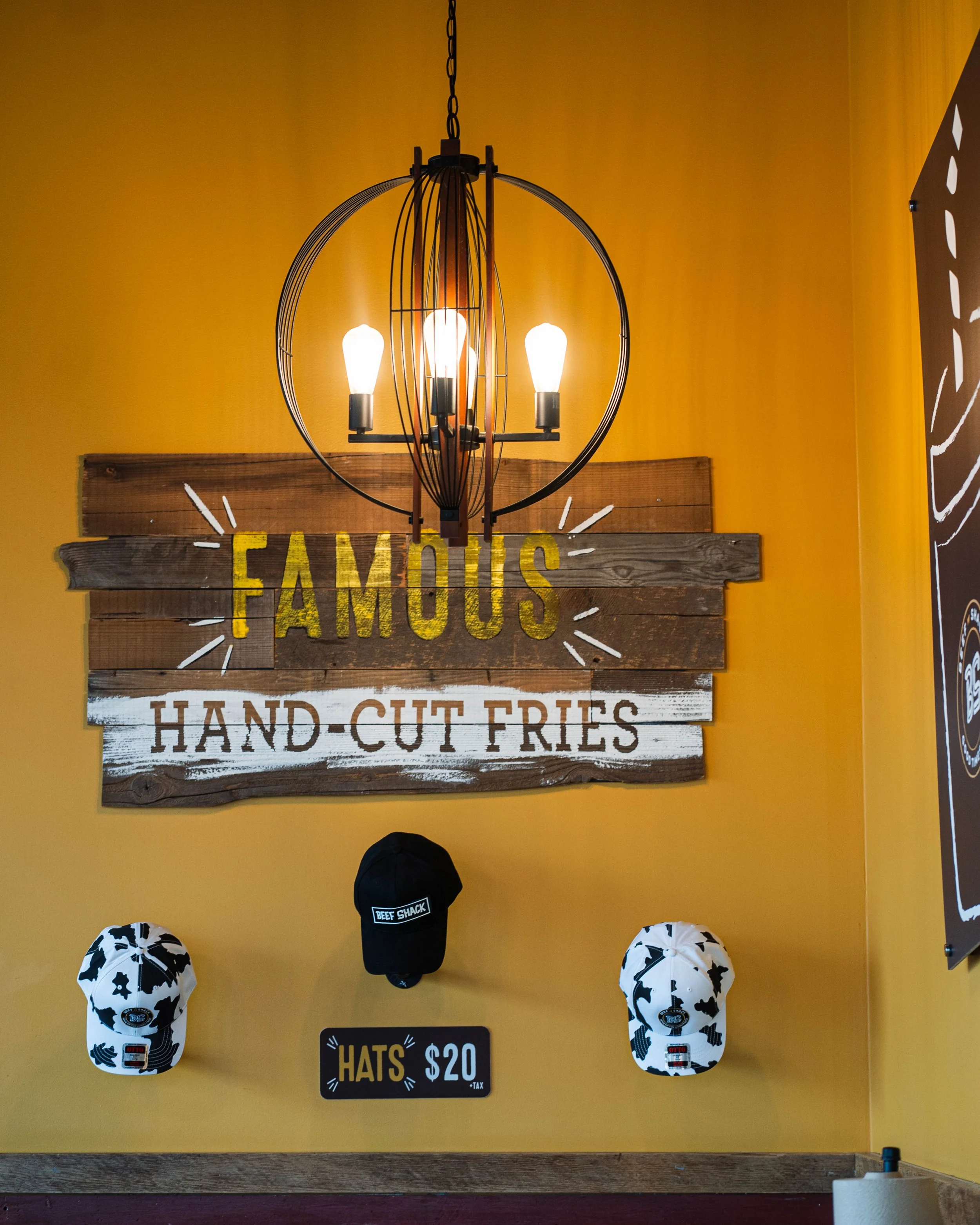 Decorative restaurant interior with a yellow wall, wooden sign reading 'FAMOUS HAND-CUT FRIES,' hanging black hat with 'BEEF SHACK' logo, and two cow-patterned hats, illuminated by a modern hanging light fixture.