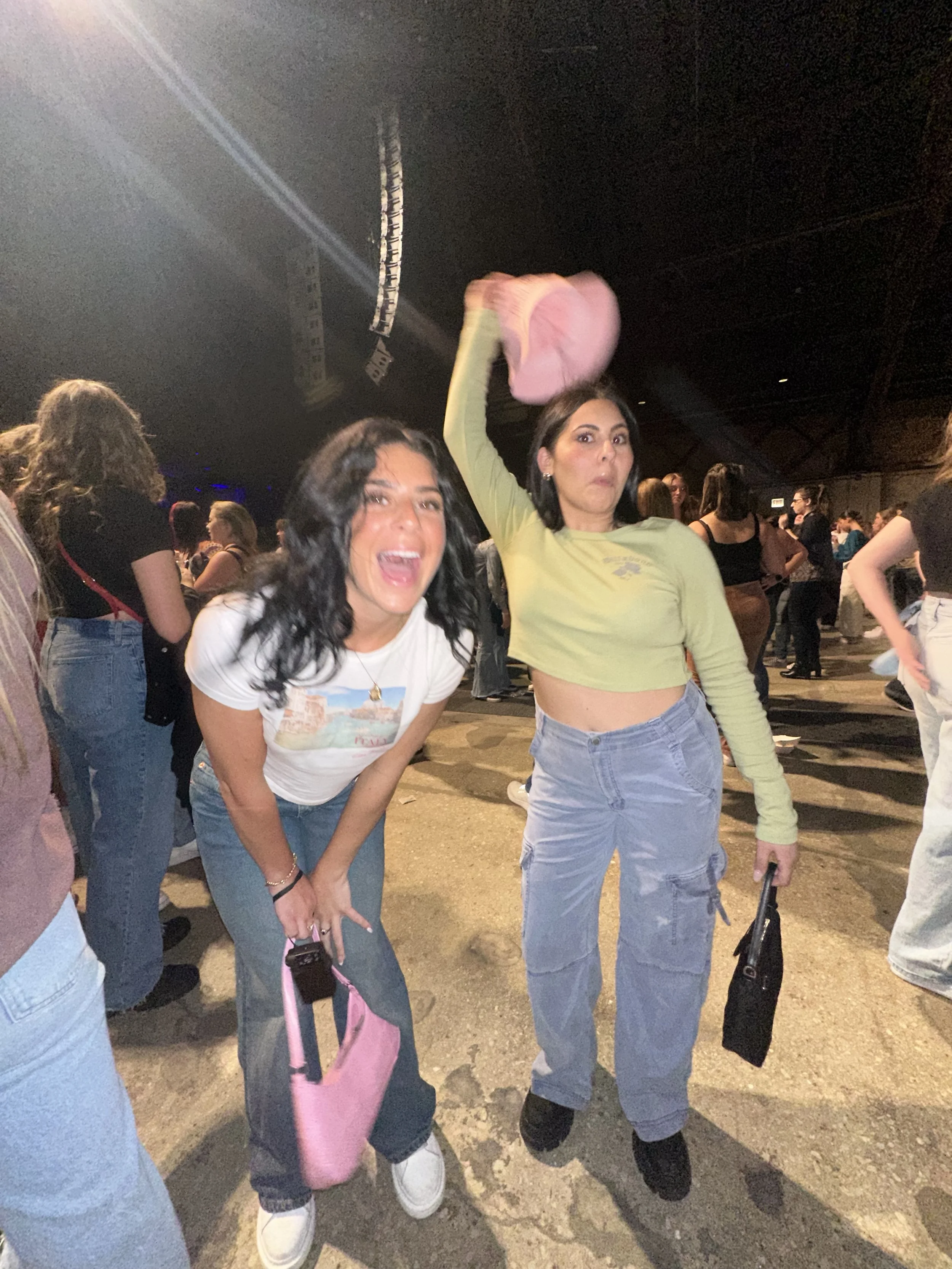 Two women at a crowded indoor event, one is smiling and the other appears surprised, holding a pink bag and a black bag respectively.