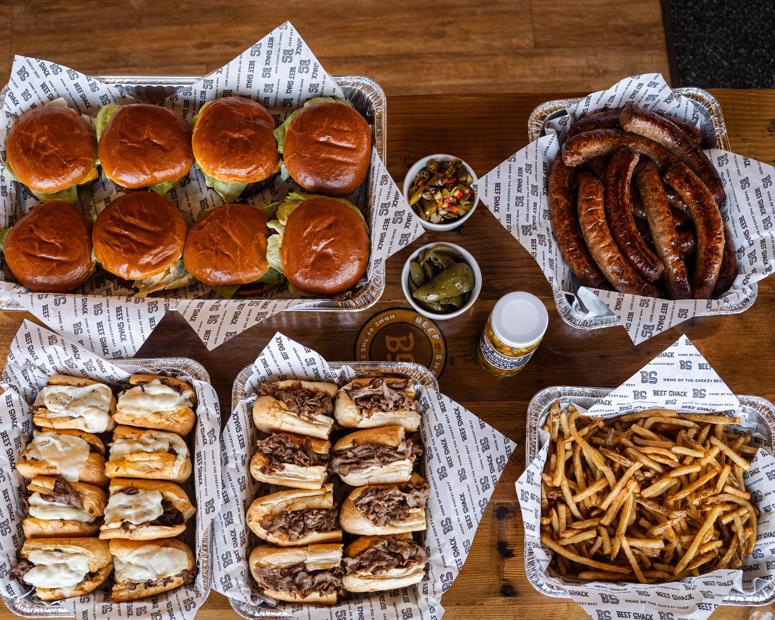 An array of barbecue foods including sliders, hot dogs, sausages, French fries, and mozzarella sticks, along with small cups of pickles and jalapeños, served on a wooden table.