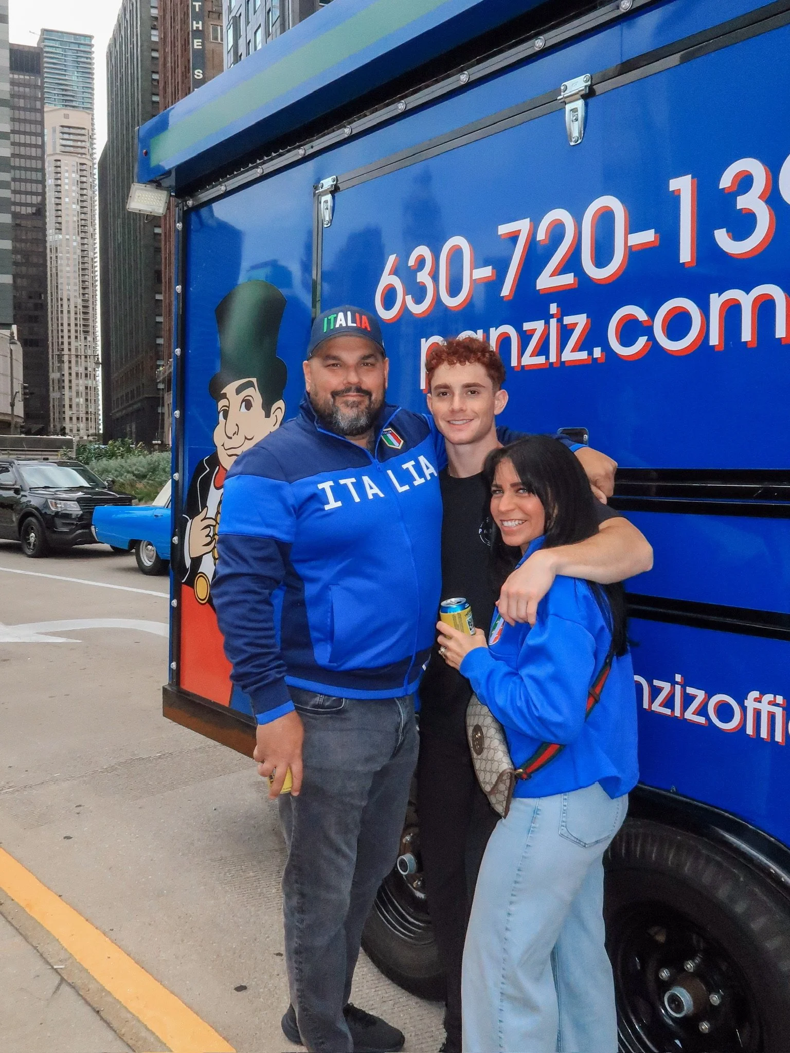 Three people standing in front of a blue food truck with Italian-themed graphics and text, including a cartoon chef, phone number, and website. They are smiling, with two of them wearing blue Italy jackets and one holding a beverage can.