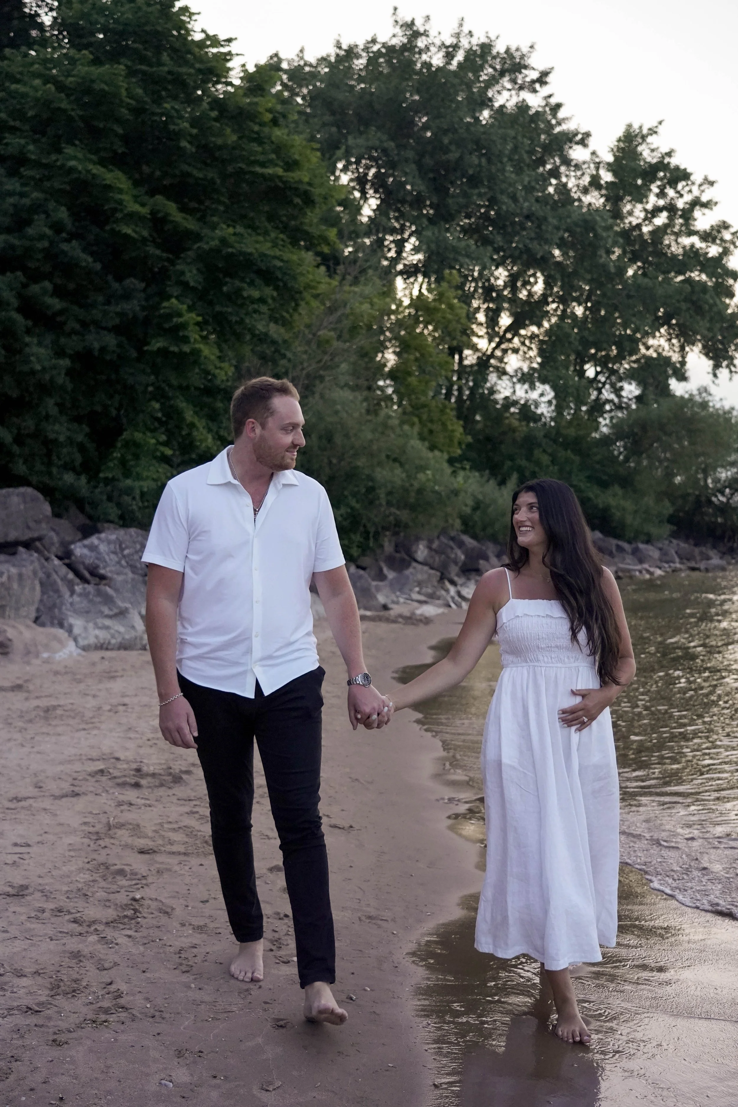 A couple walks hand in hand along a beach at sunset, with the woman holding her stomach and smiling, and the man looking at her affectionately. The scene has trees and rocks in the background.