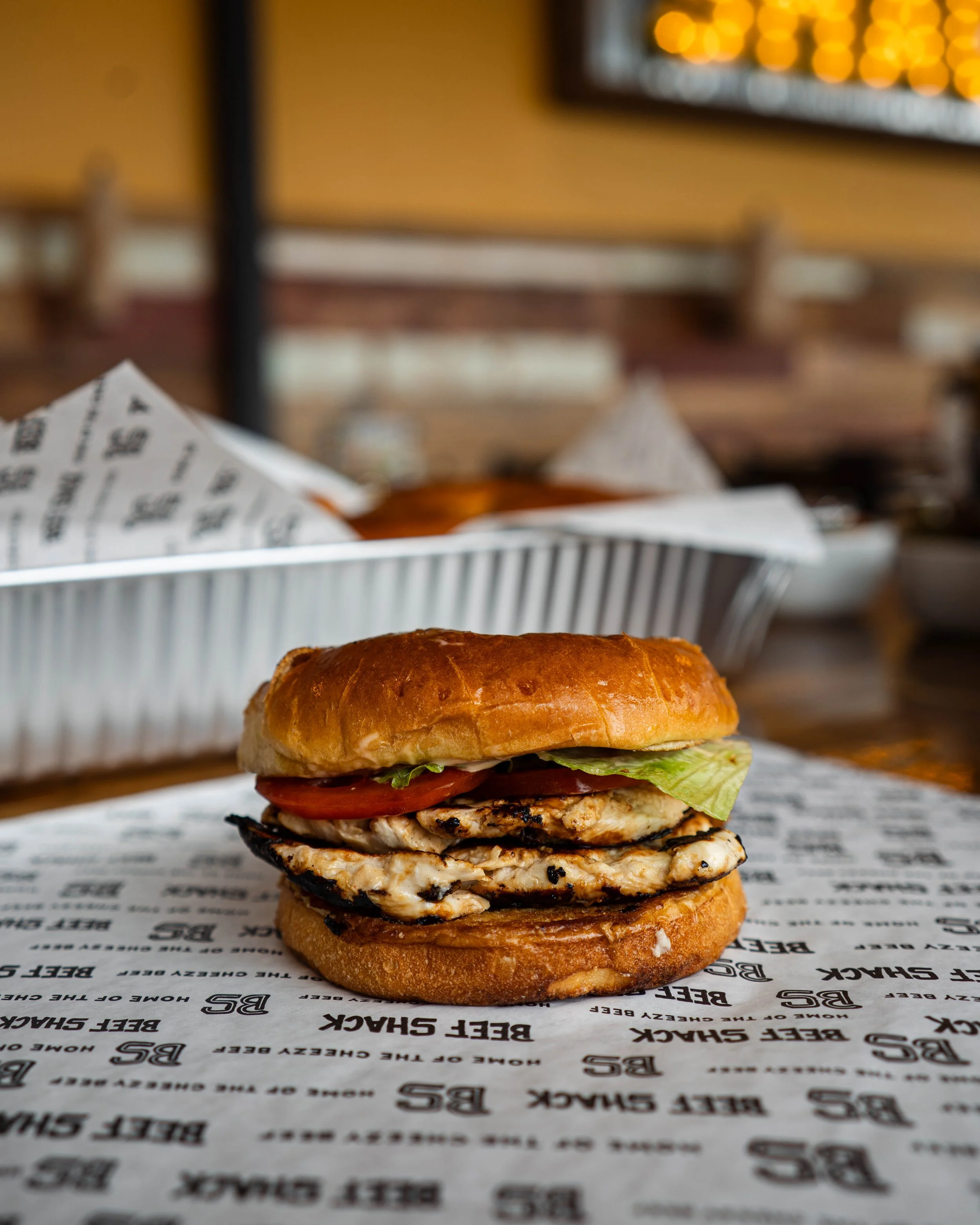 A grilled chicken sandwich with lettuce, tomato, and grilled onions on a bun, placed on branded paper, with a metal basket of fries in the background, inside a casual restaurant.