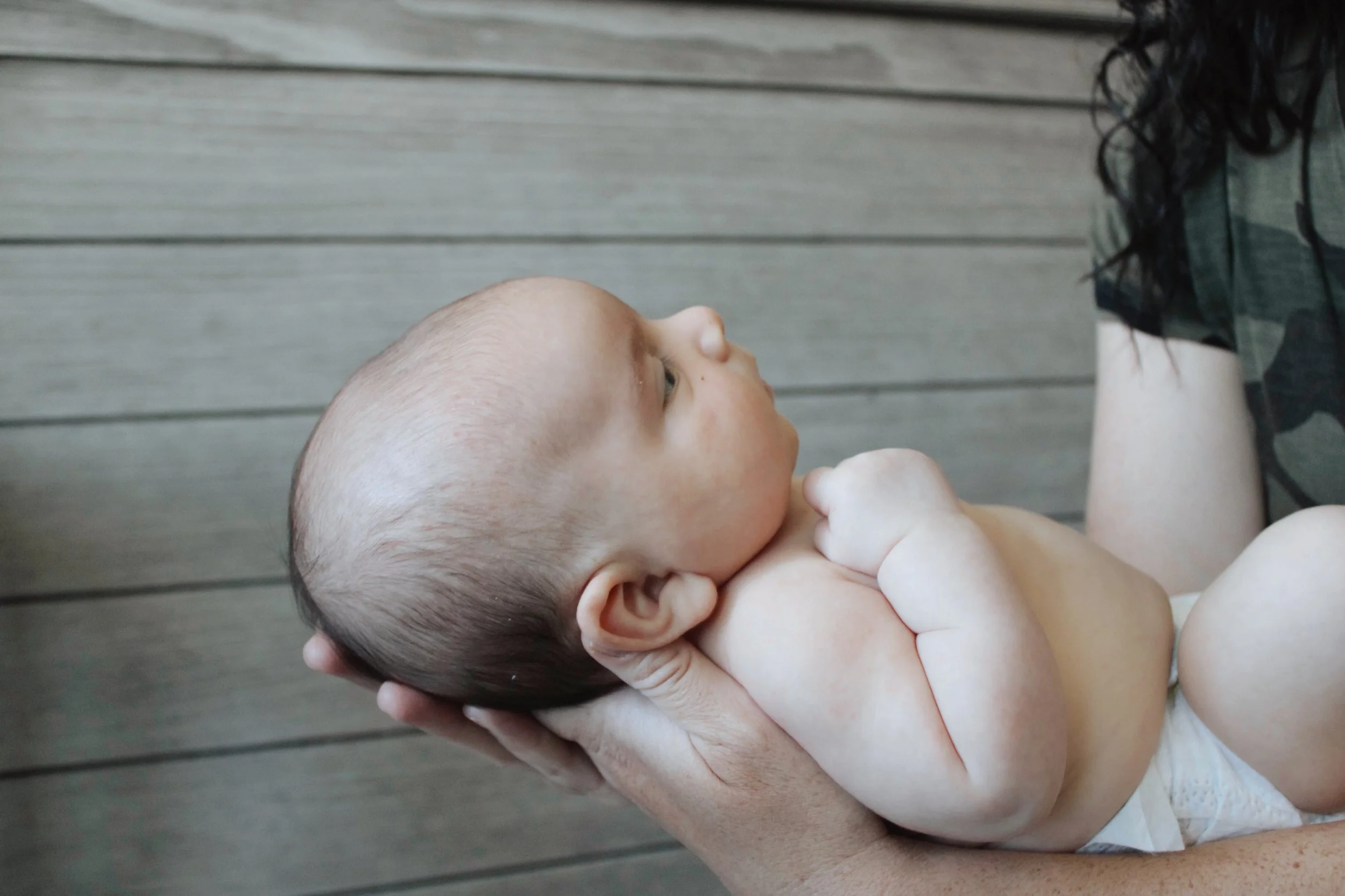 A baby with light skin and a bald head lying in an adult's arms, looking to the side, with a wooden background.