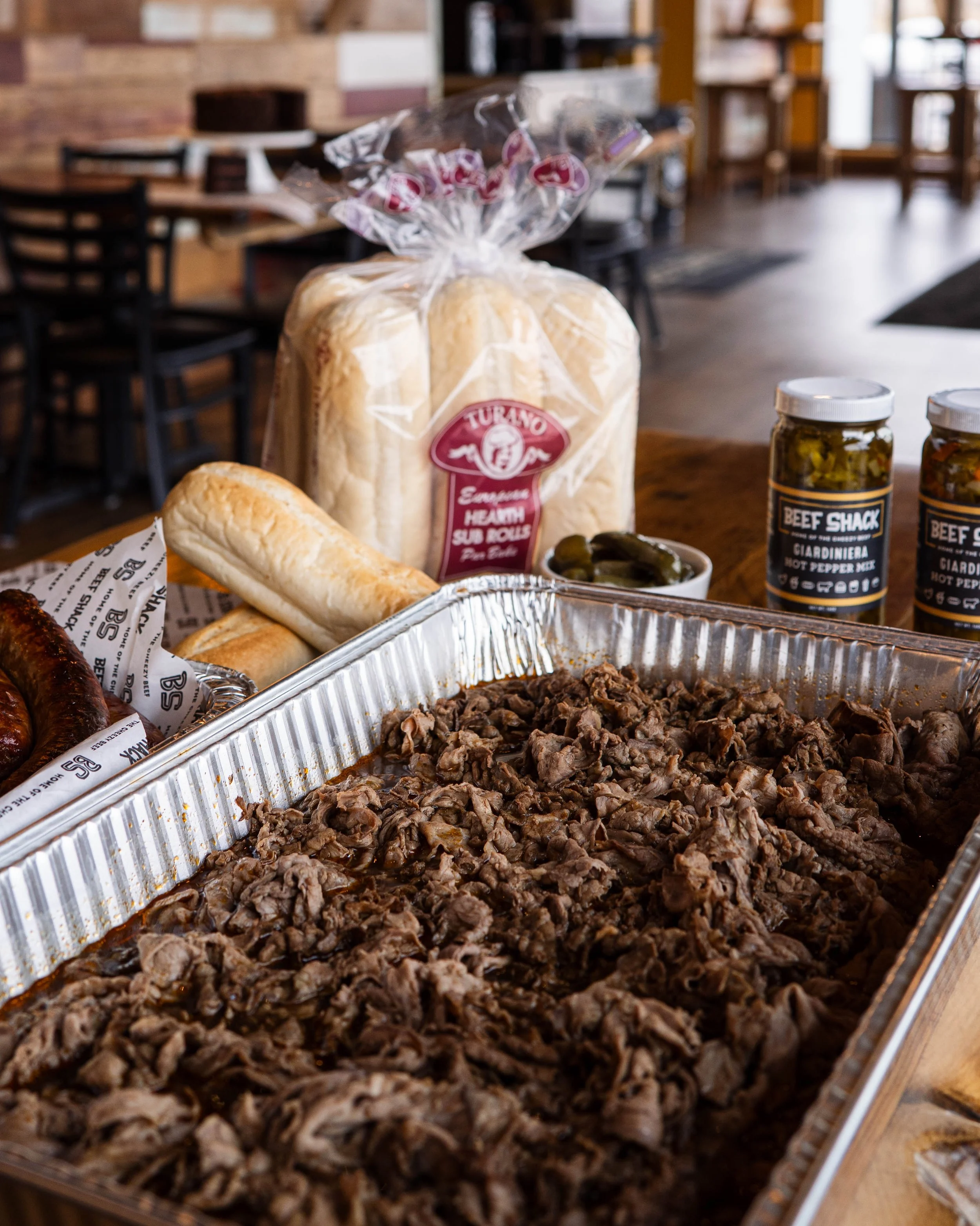 Aluminum tray filled with shredded cooked beef, with bread rolls, pickles, and condiments in jars and packaged bread on a wooden table in a restaurant setting.