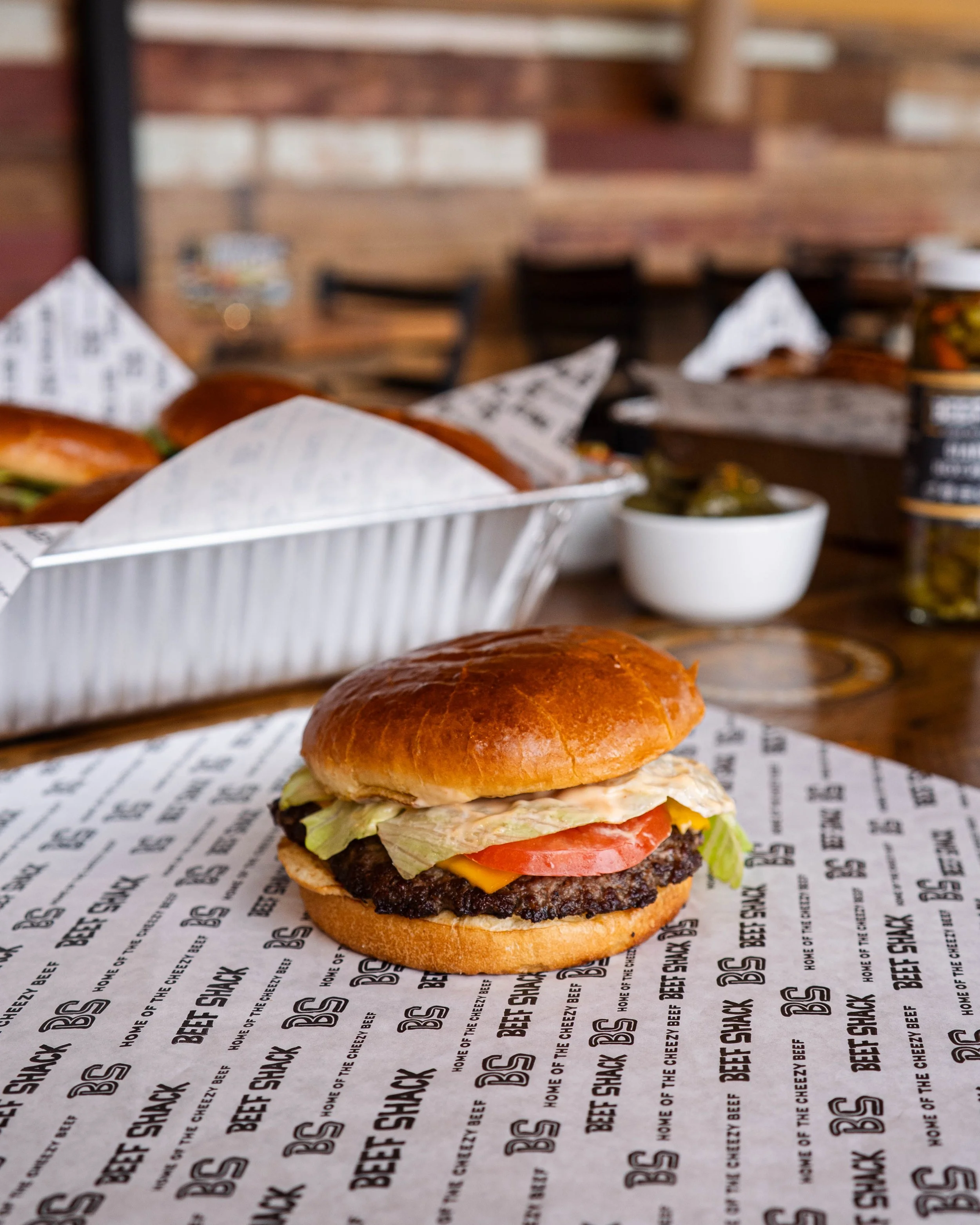Close-up of a cheeseburger with lettuce, tomato, and pickles in a bun on branded paper at a restaurant table. In the background, there are more burgers, a small bowl of pickles, and condiments on a wooden table.