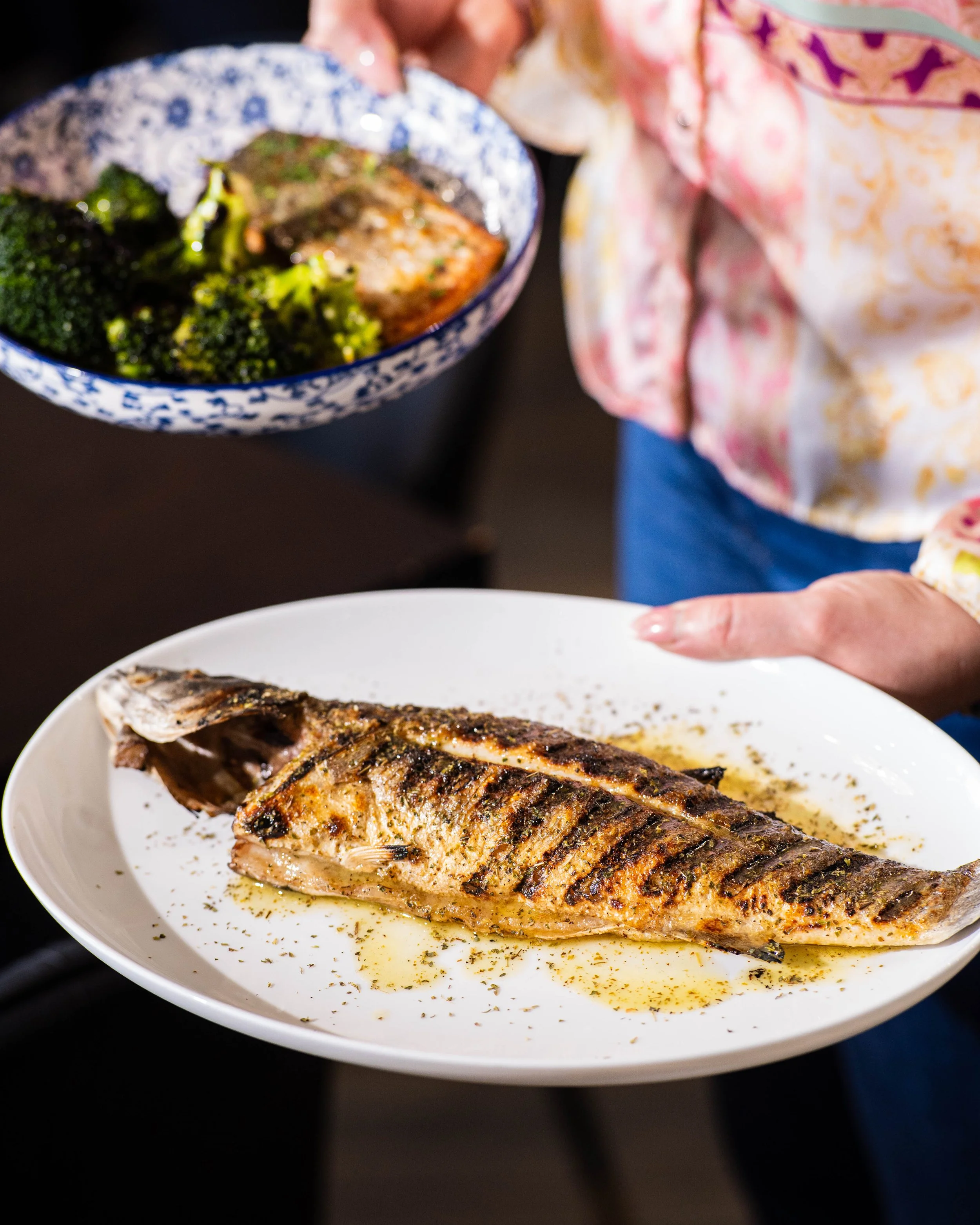 Person holding a white plate with grilled fish garnished with herbs and oil, and another person showing a bowl of cooked broccoli and baked fish.