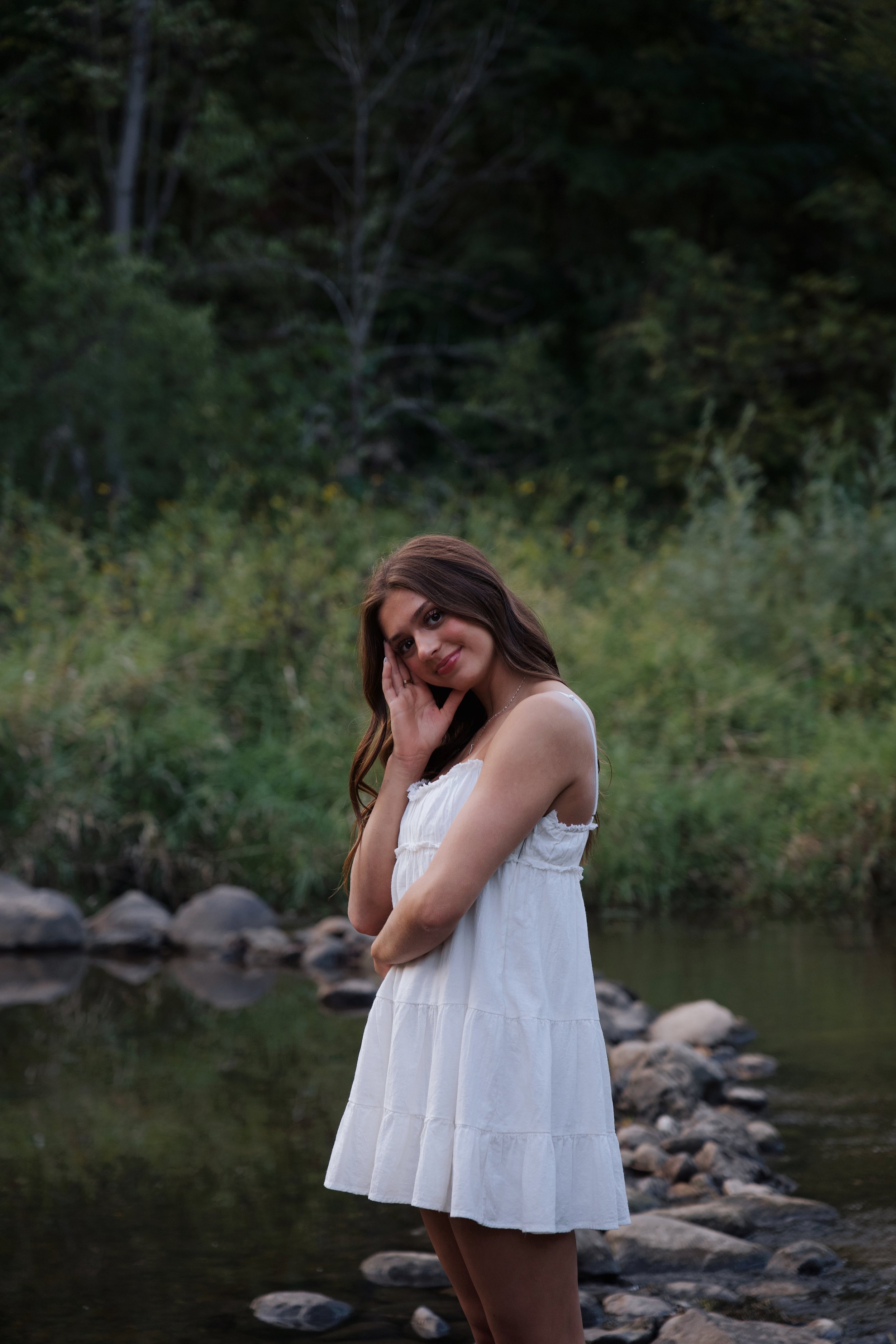 A young woman wearing a white dress standing by a creek in a forest setting, smiling and posing with one hand touching her face.