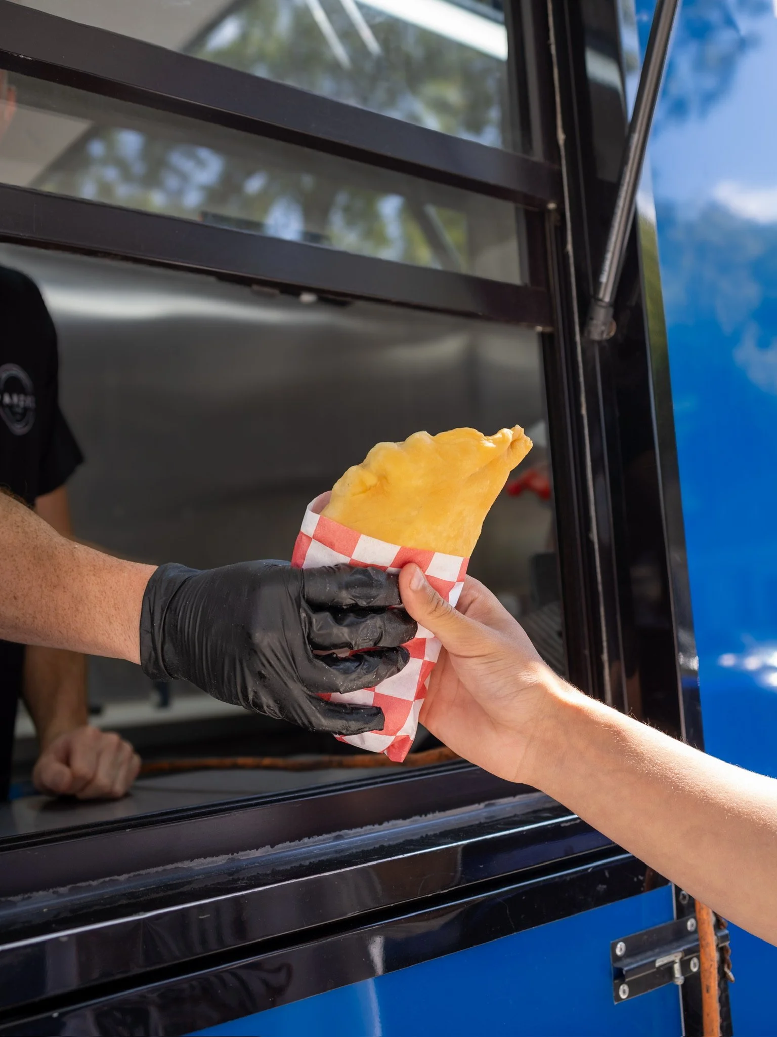 A person in a black glove is handing a fried snack in a checkered paper wrapper to another person outside a blue food truck with a window.