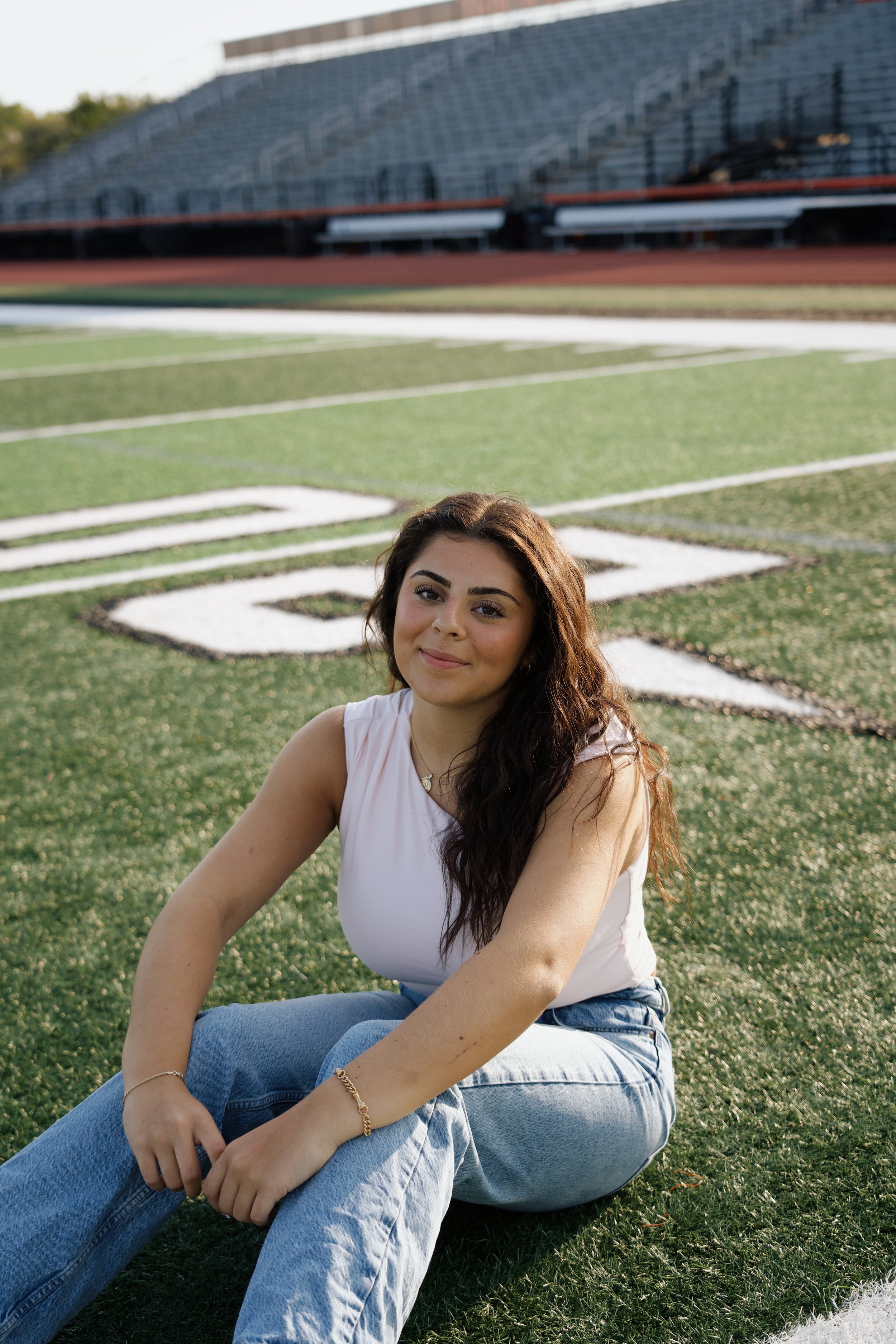 Young woman with long dark hair sitting on a green football field with stadium bleachers in the background.
