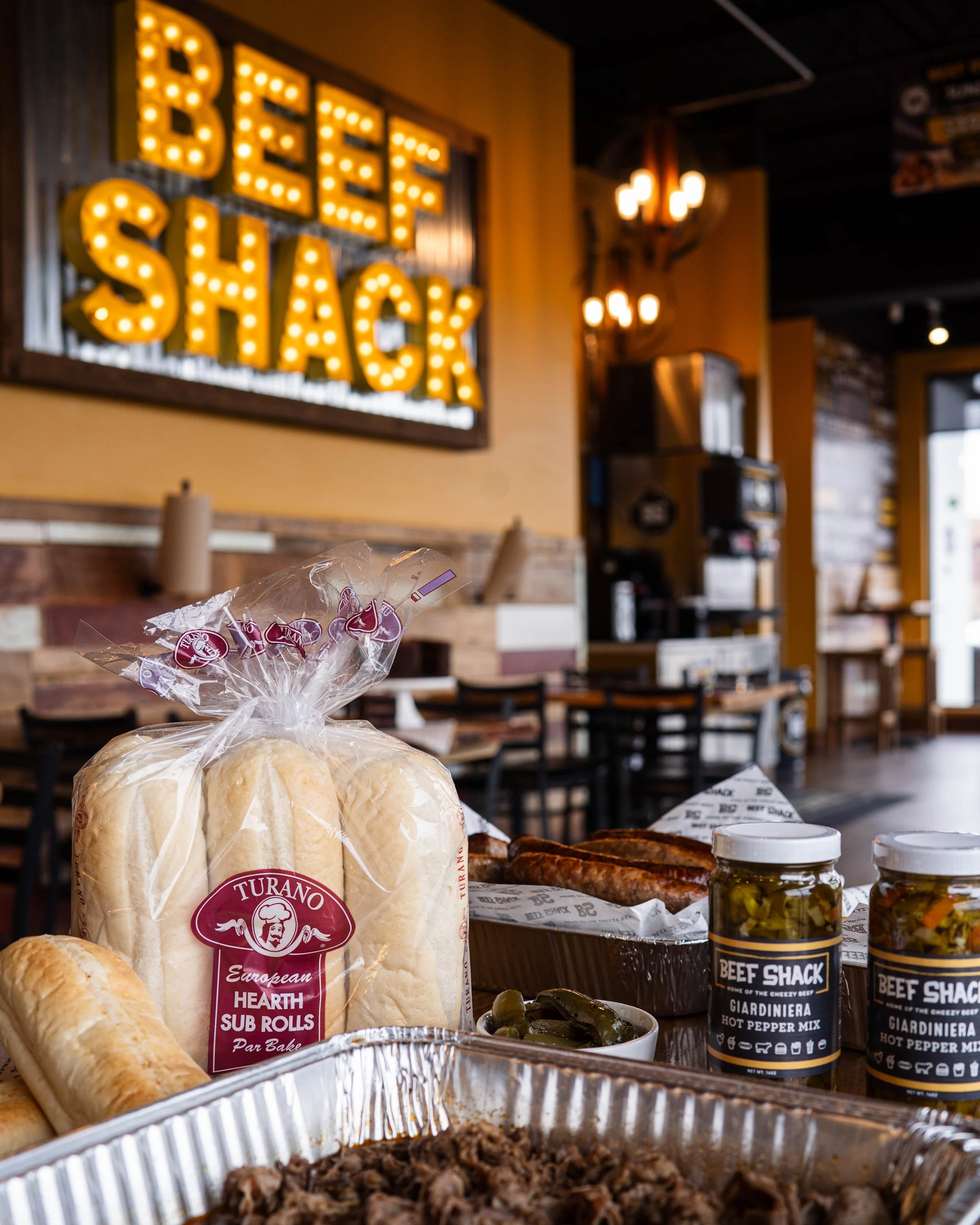 Inside a restaurant called Beef Shack, with a glowing sign on the wall, showing various meats, bread, hot peppers, and condiments on the table.