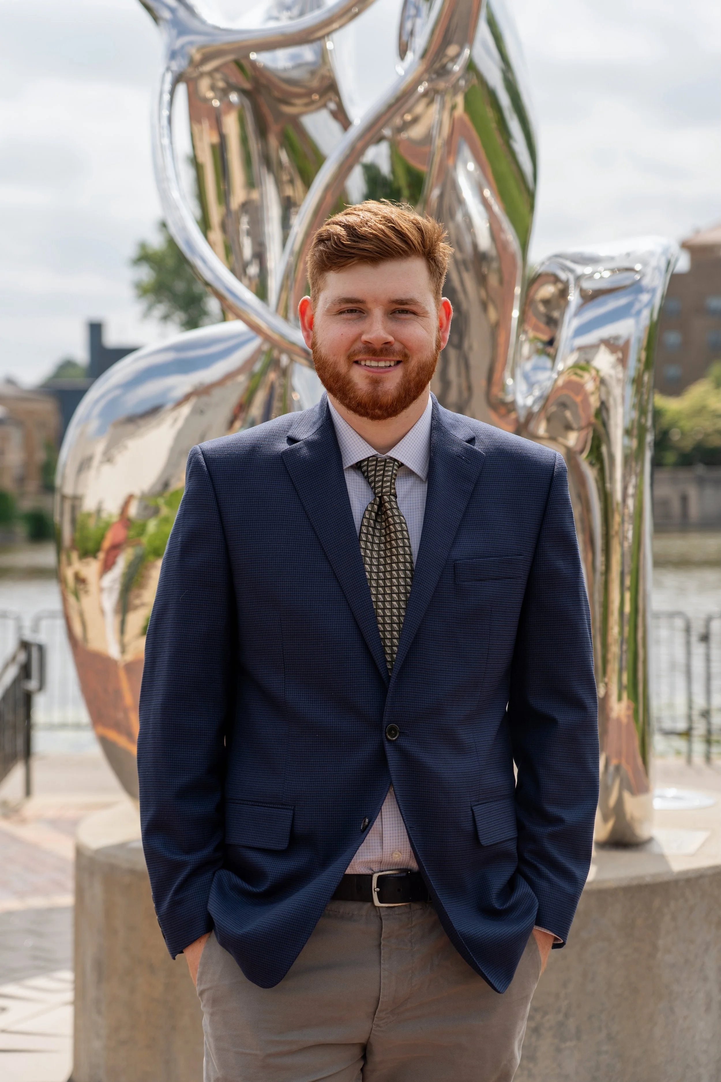 A man in a navy blazer, white shirt, patterned tie, and khaki pants smiling outdoors with a large metallic sculpture and river in the background.