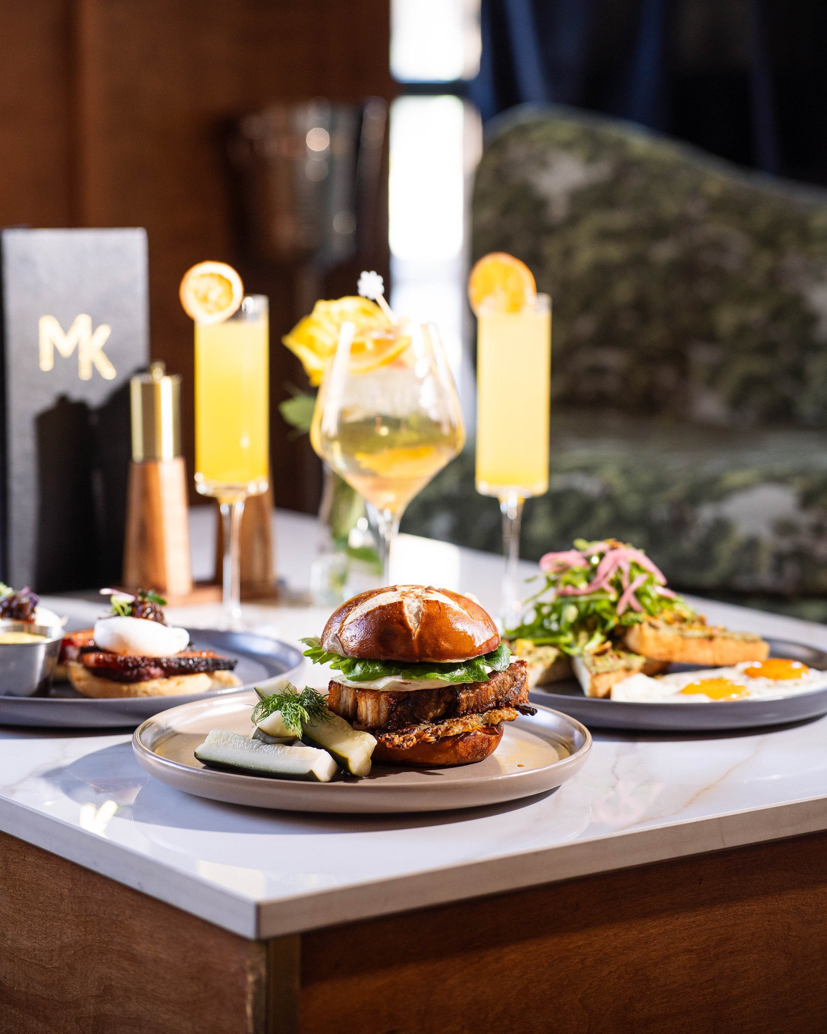 Three cocktails with citrus garnishes, a burger with lettuce, bacon, and pickles, fried eggs with ham and greens, and other small dishes on a marble counter.