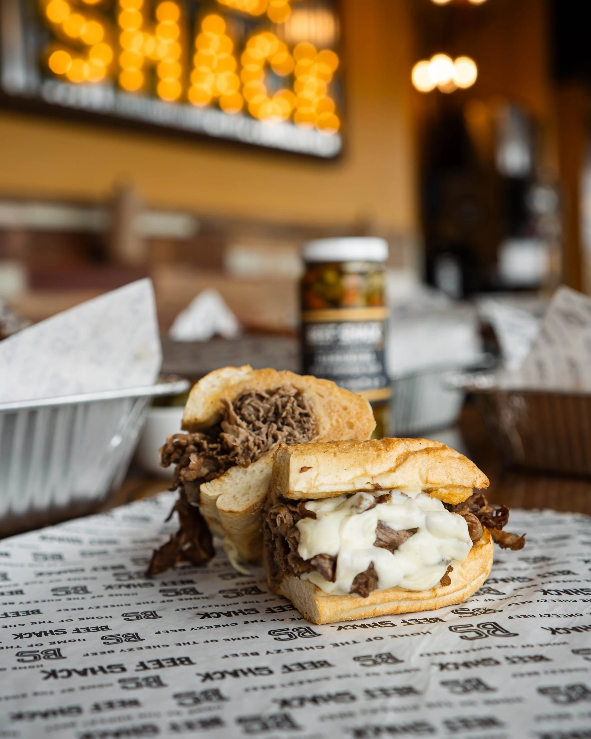 A burger with sliced beef, melted cheese, and a bun in a restaurant setting, with a blurred background and a paper wrap that says 'Beef Shack'.