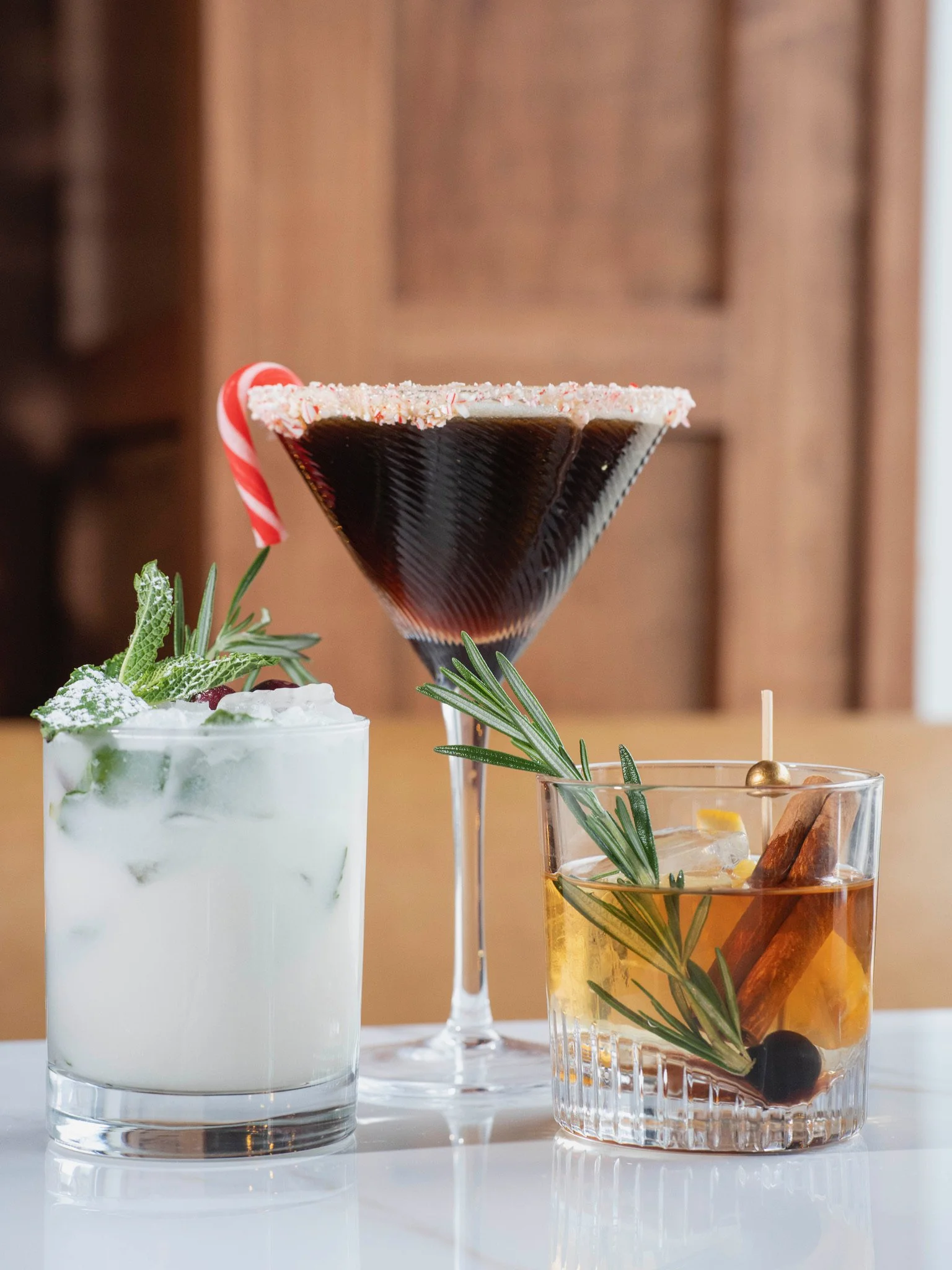 Three holiday cocktails on a white surface with a wood background: a creamy white drink garnished with mint and sprigs, a large martini glass with a dark cocktail and a sugared rim with a peppermint candy, and a bourbon or whiskey drink with a cinnam