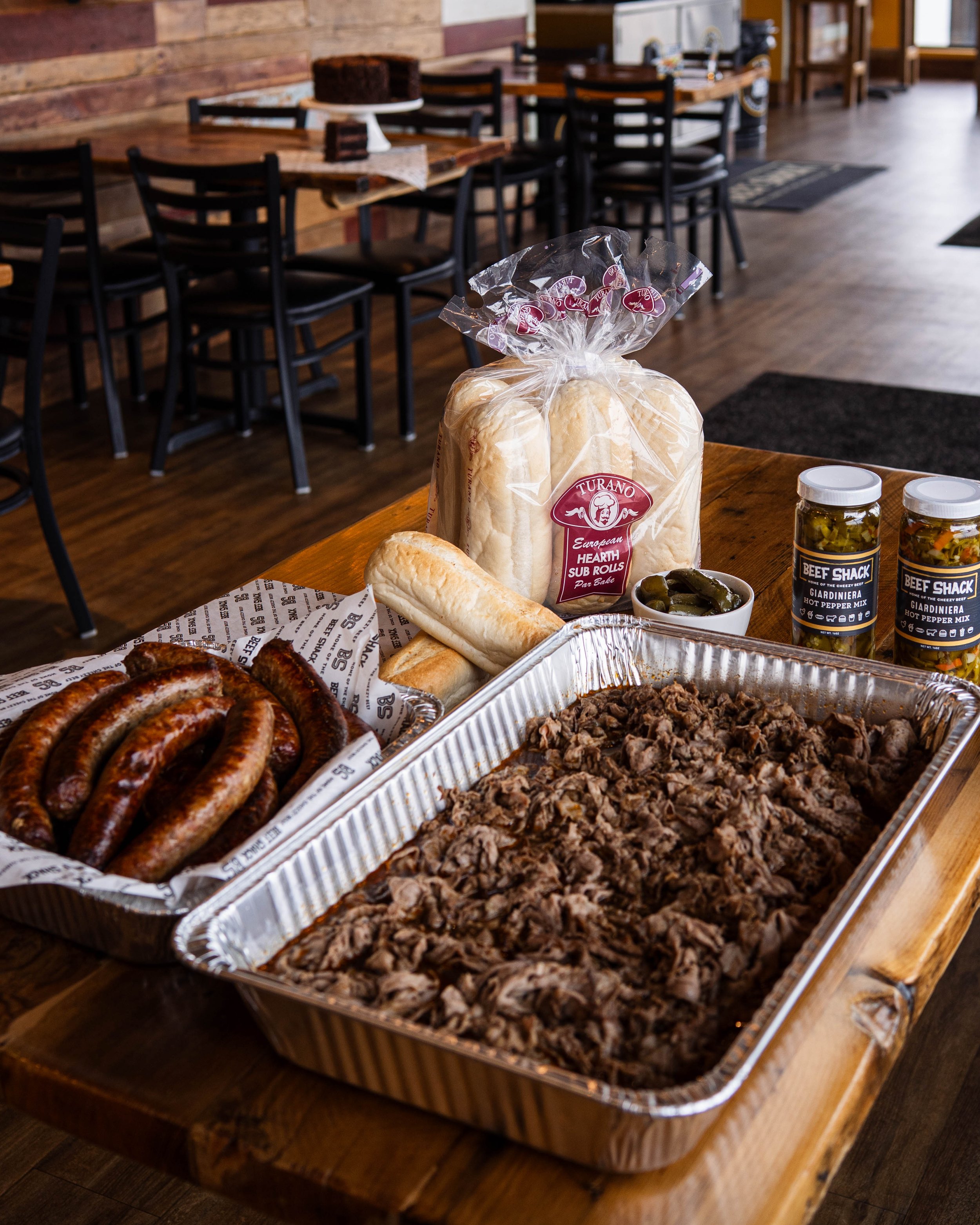 Barbecue food arrangement on a wooden table, including cooked sausages, shredded beef, bread rolls, pickles, and condiments in jars, with a bakery loaf of bread in the background inside a restaurant.
