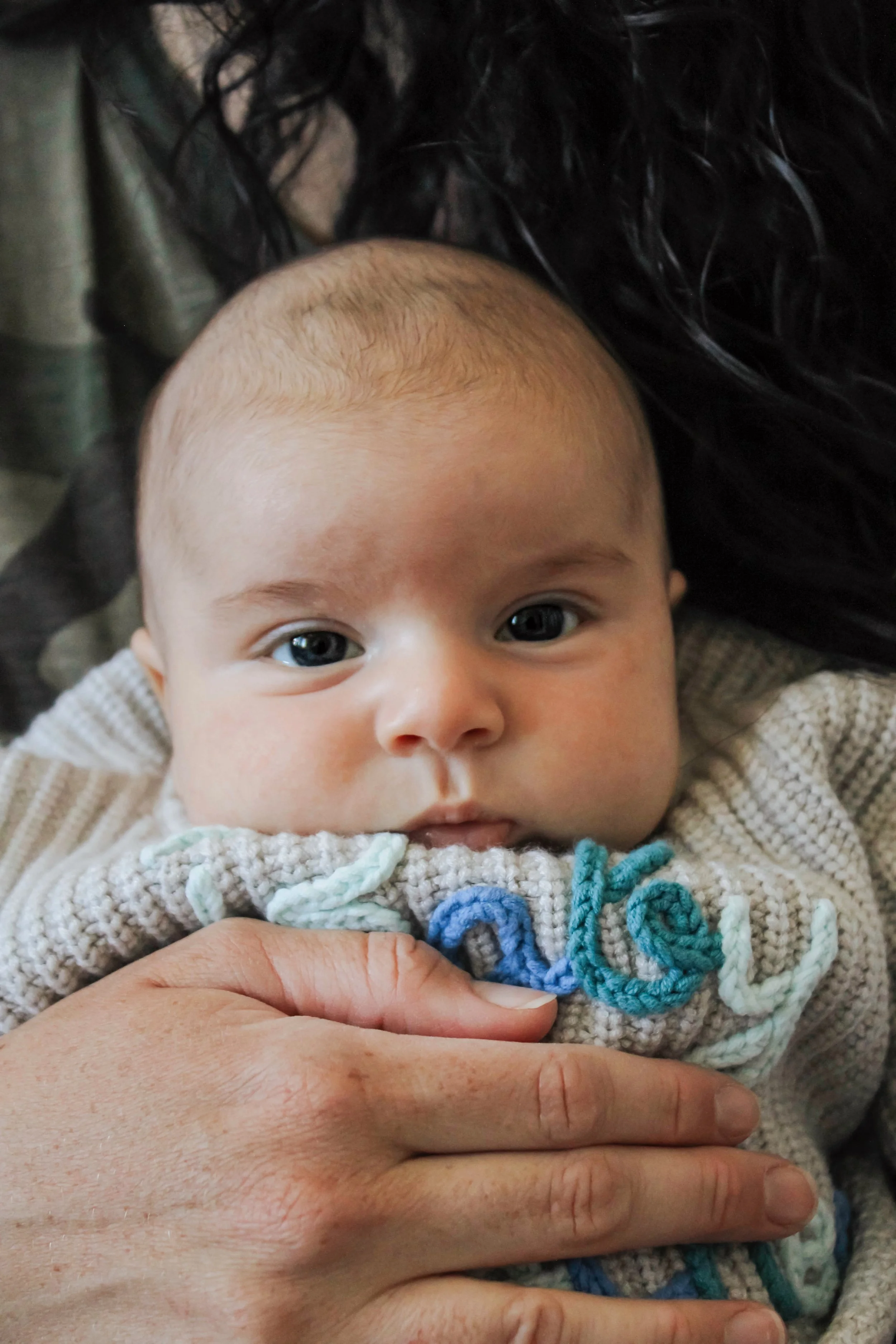 Close-up of a baby with blue eyes and light skin, lying down, wearing a beige knitted sweater with the word "love" embroidered in colorful thread, held gently by an adult's hand.
