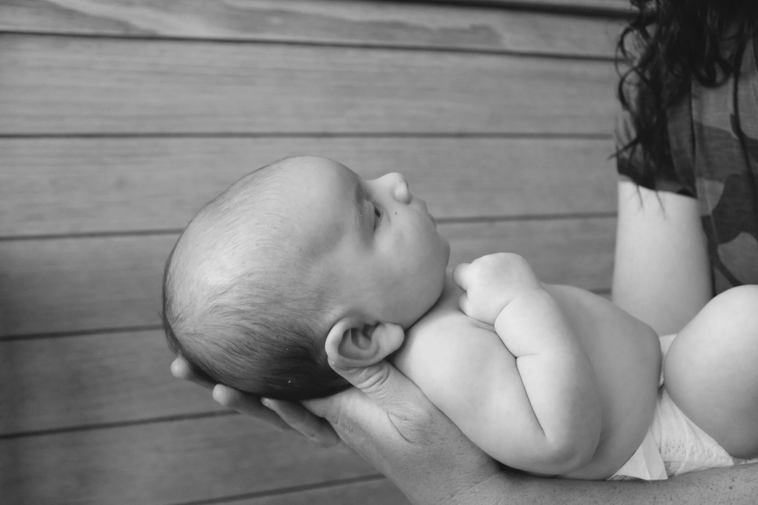A baby with a shaved head being gently held by an adult. The baby is looking up at the adult with a wooden wall in the background.