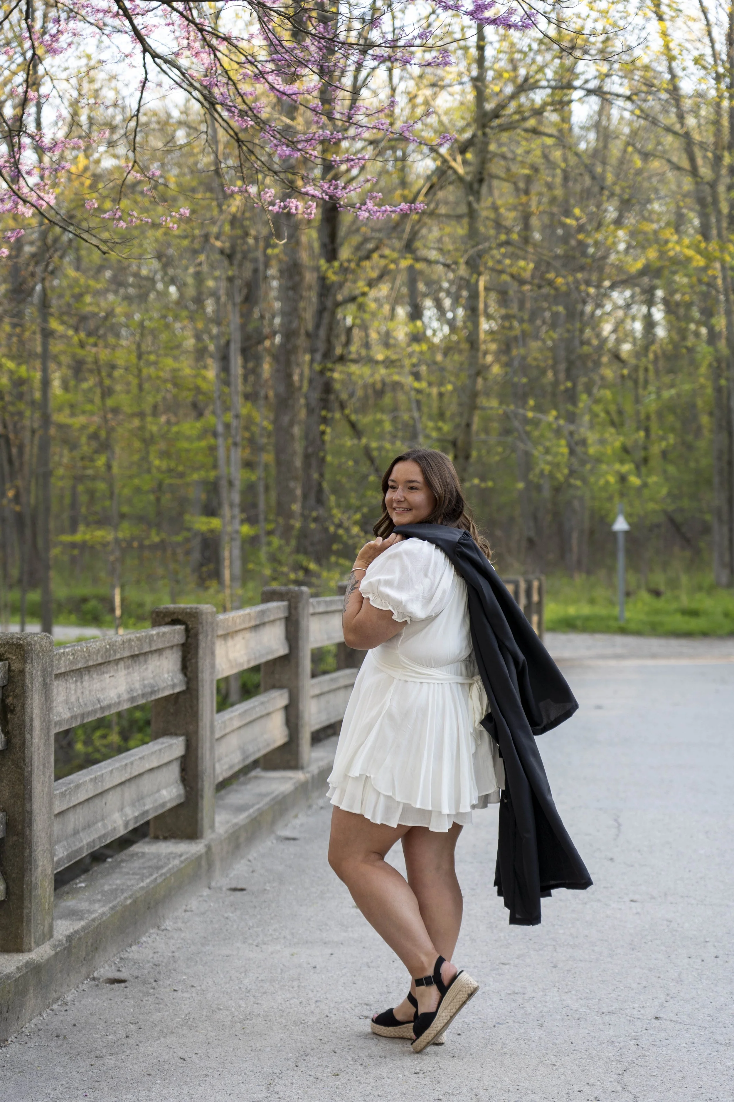 A woman in a white dress standing on a sidewalk near a wooden fence, holding a black jacket over her shoulder, smiling, with trees and pink blossoms in the background.