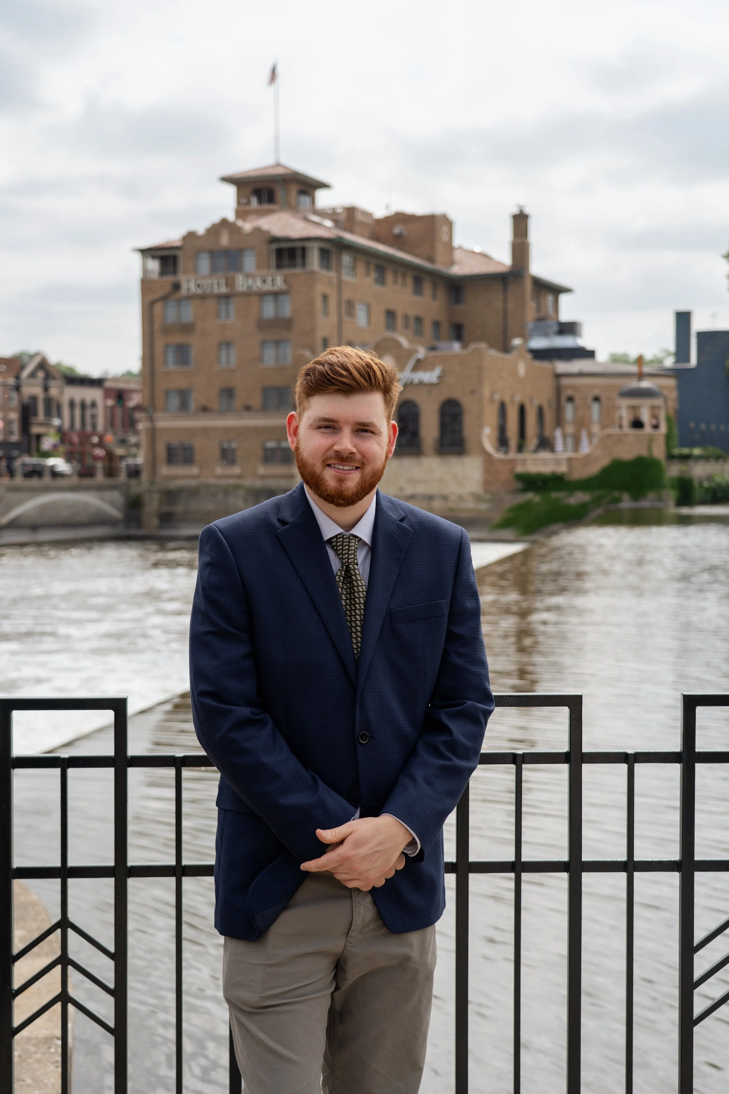 A young man with red hair and beard wearing a navy blazer, dress shirt, and tie, standing by a river with a historic hotel building in the background.
