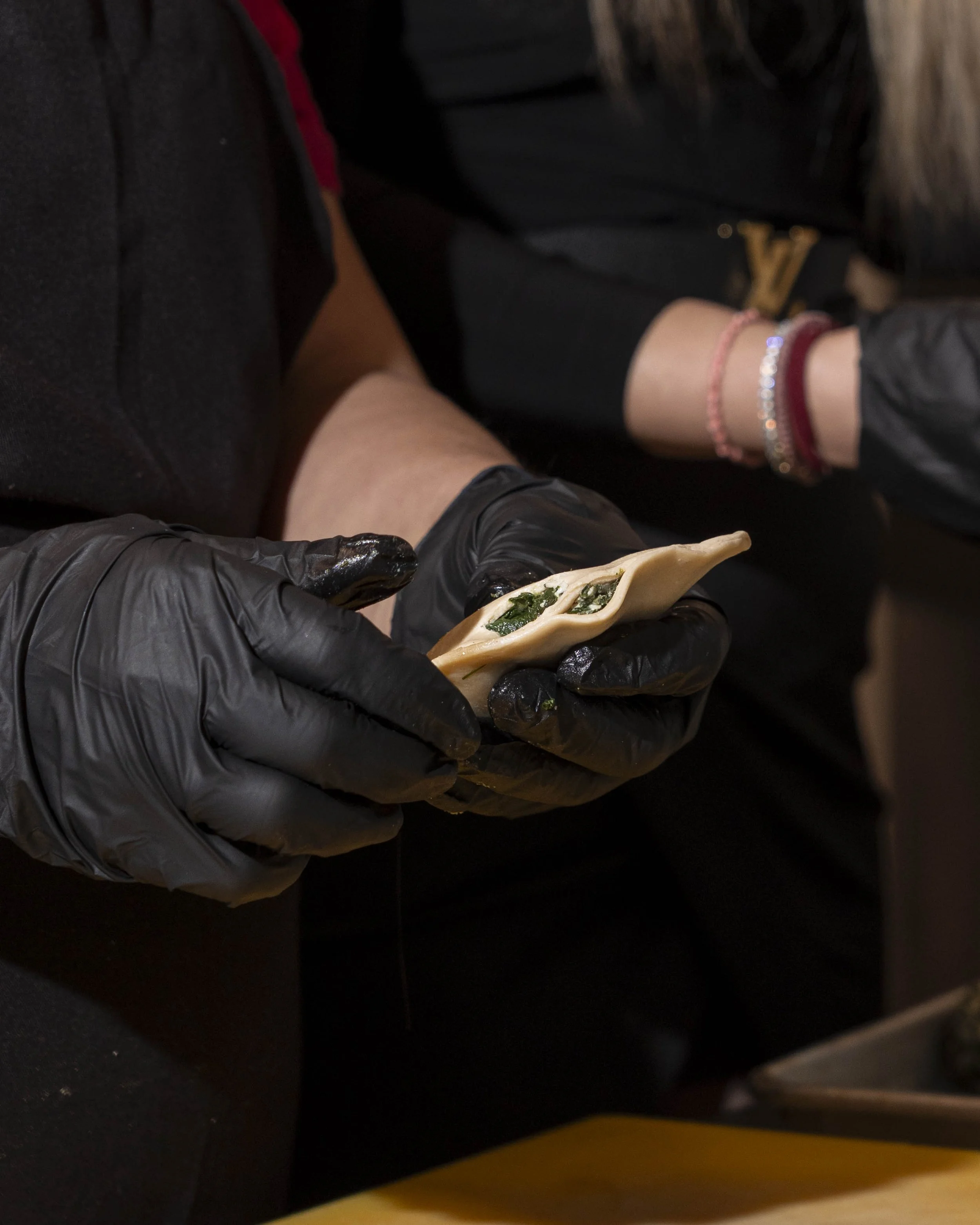 A person wearing black gloves holding a half-moon shaped dumpling filled with green vegetables.