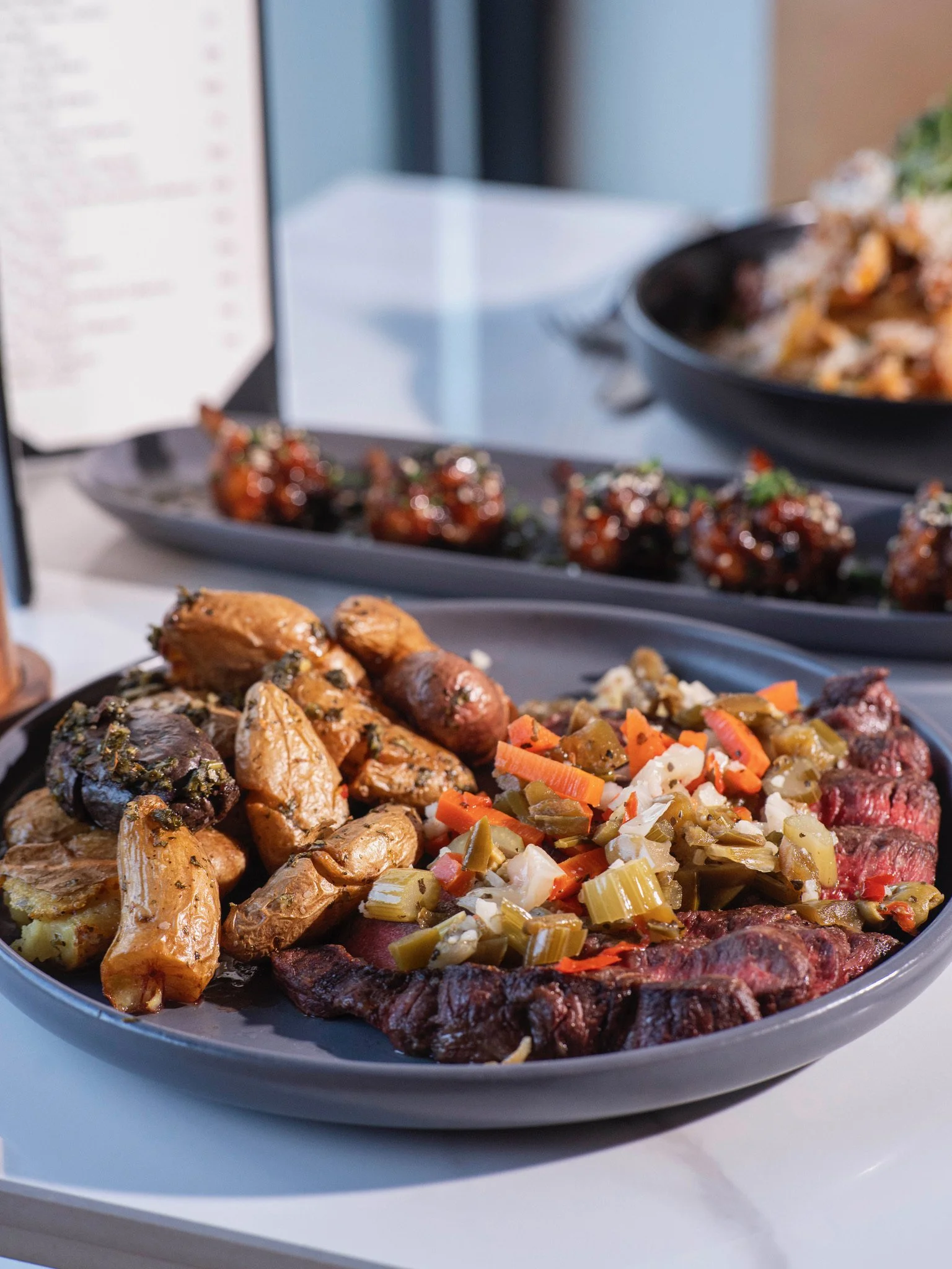 A plate of roasted vegetables and sliced cooked meat, topped with chopped vegetables, with other dishes in the background.
