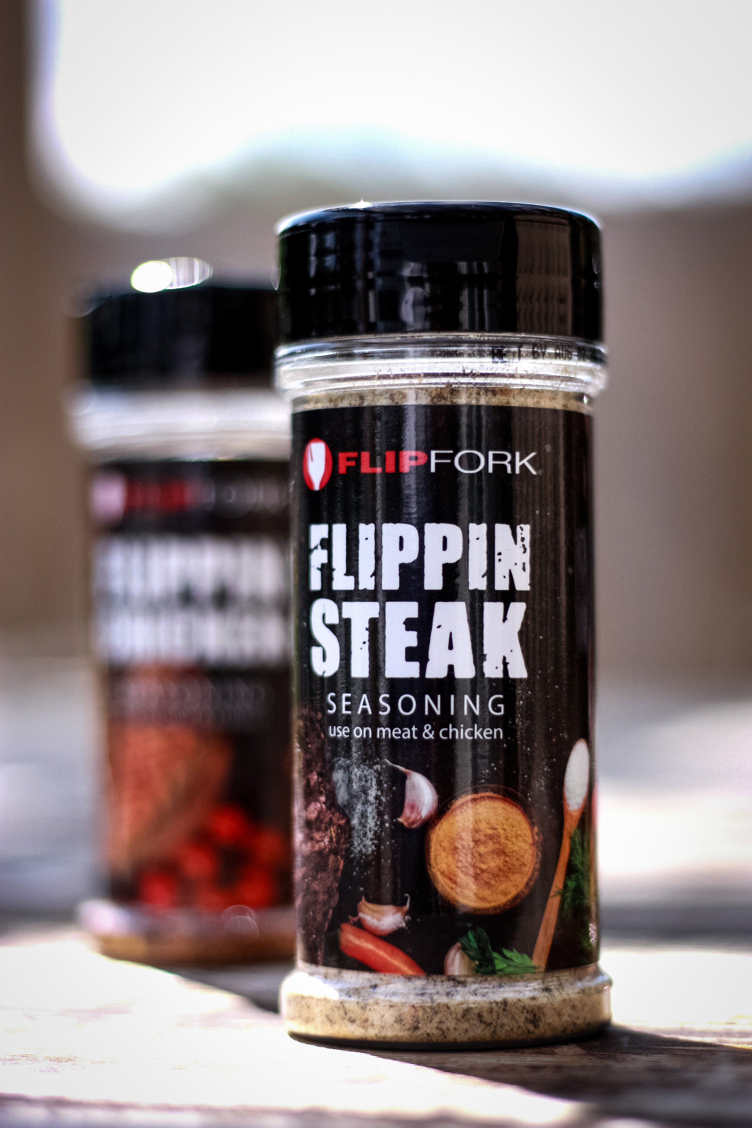 Bottle of FlipFork Flippin Steak seasoning on a wooden surface with another seasoning bottle blurred in the background.