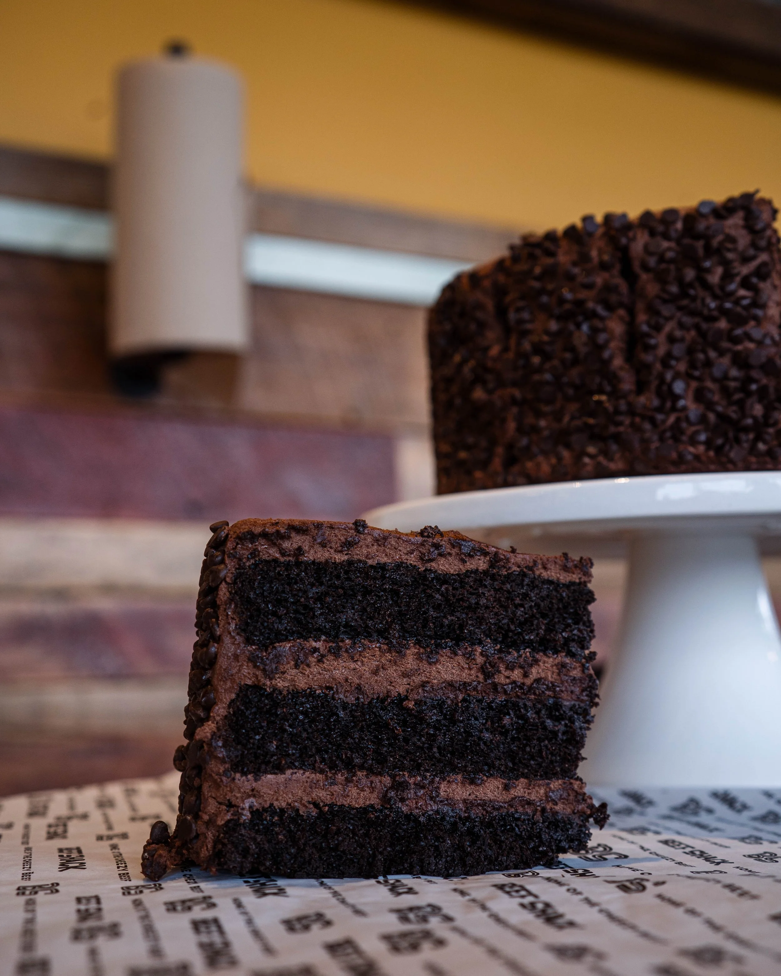A slice of chocolate cake with multiple layers and chocolate frosting, sitting on a piece of paper, with a whole chocolate cake in the background on a white cake stand.