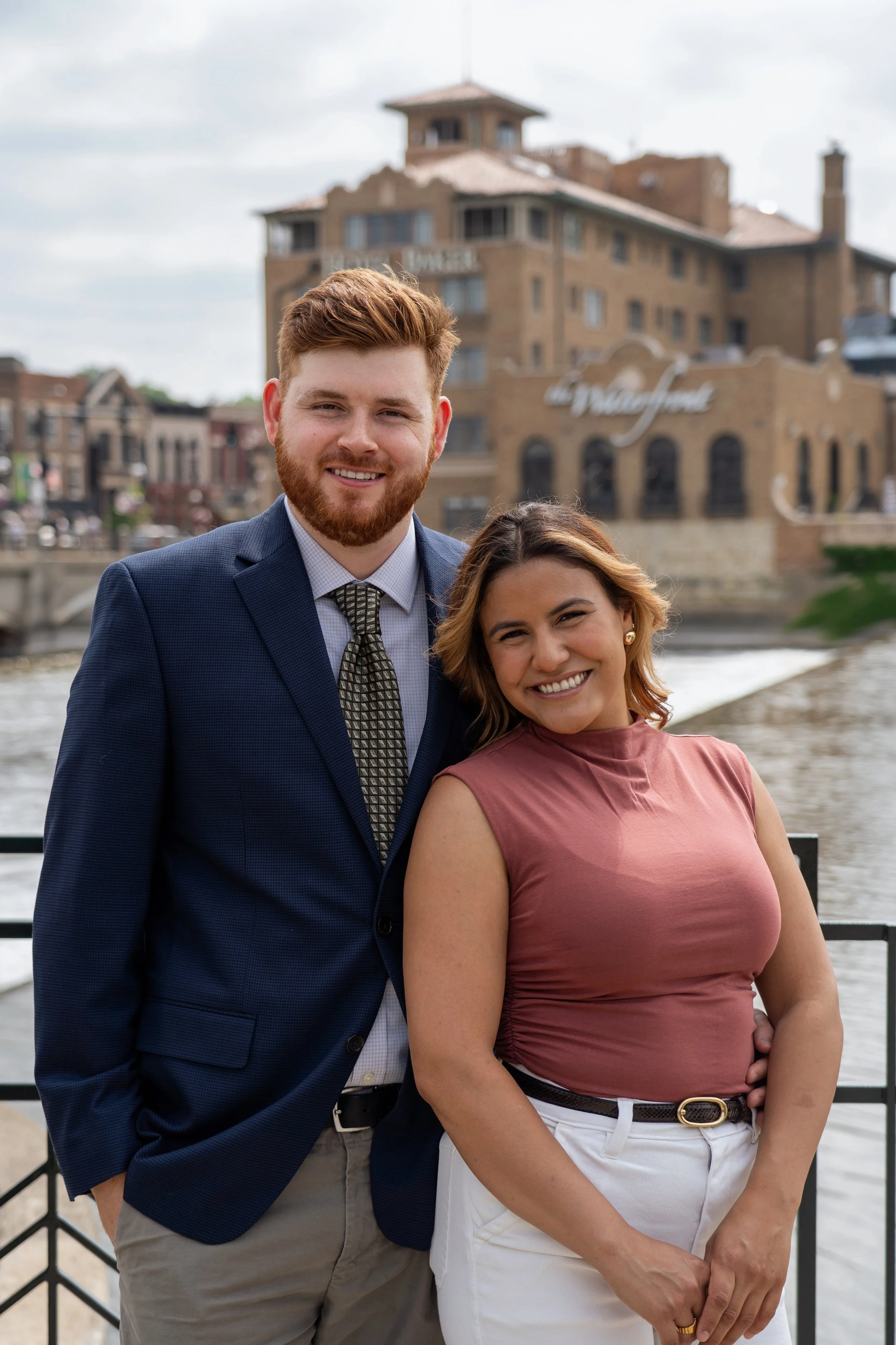 A smiling man and woman standing outdoors near a waterway, with a brick building in the background.