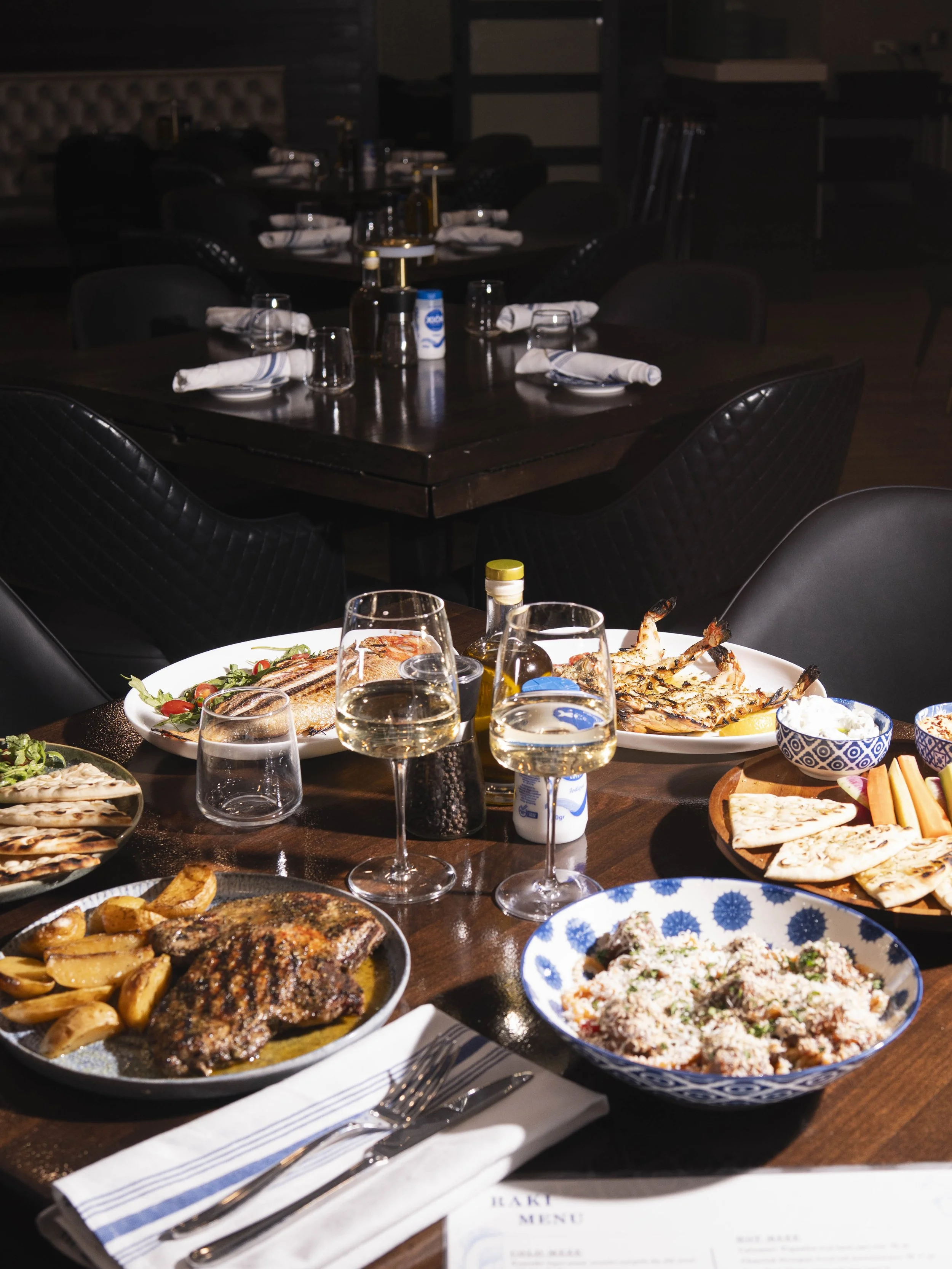 A dining table set with plates of grilled meats, shrimp, potato wedges, rice, vegetables, bread, and drinks including glasses of white wine and water, in a restaurant setting.