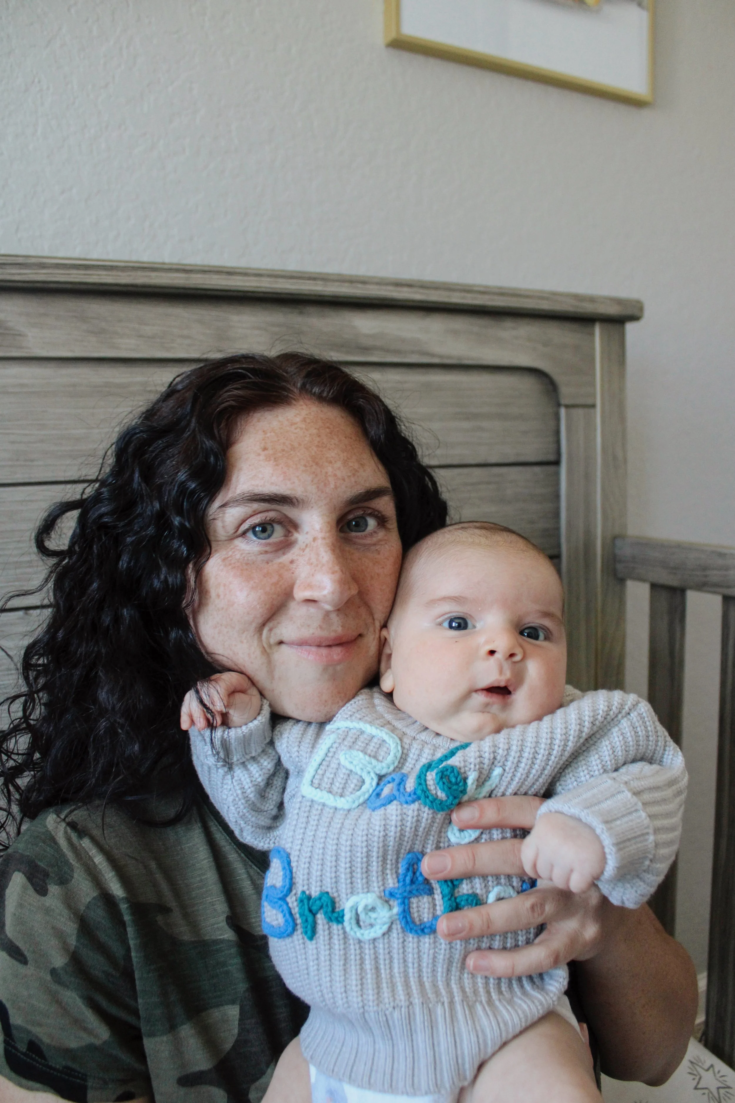 A woman with curly dark hair and freckles holding a baby with blue eyes and a surprised expression, both facing the camera indoors against a light-colored wall with a framed picture and wooden furniture in the background.