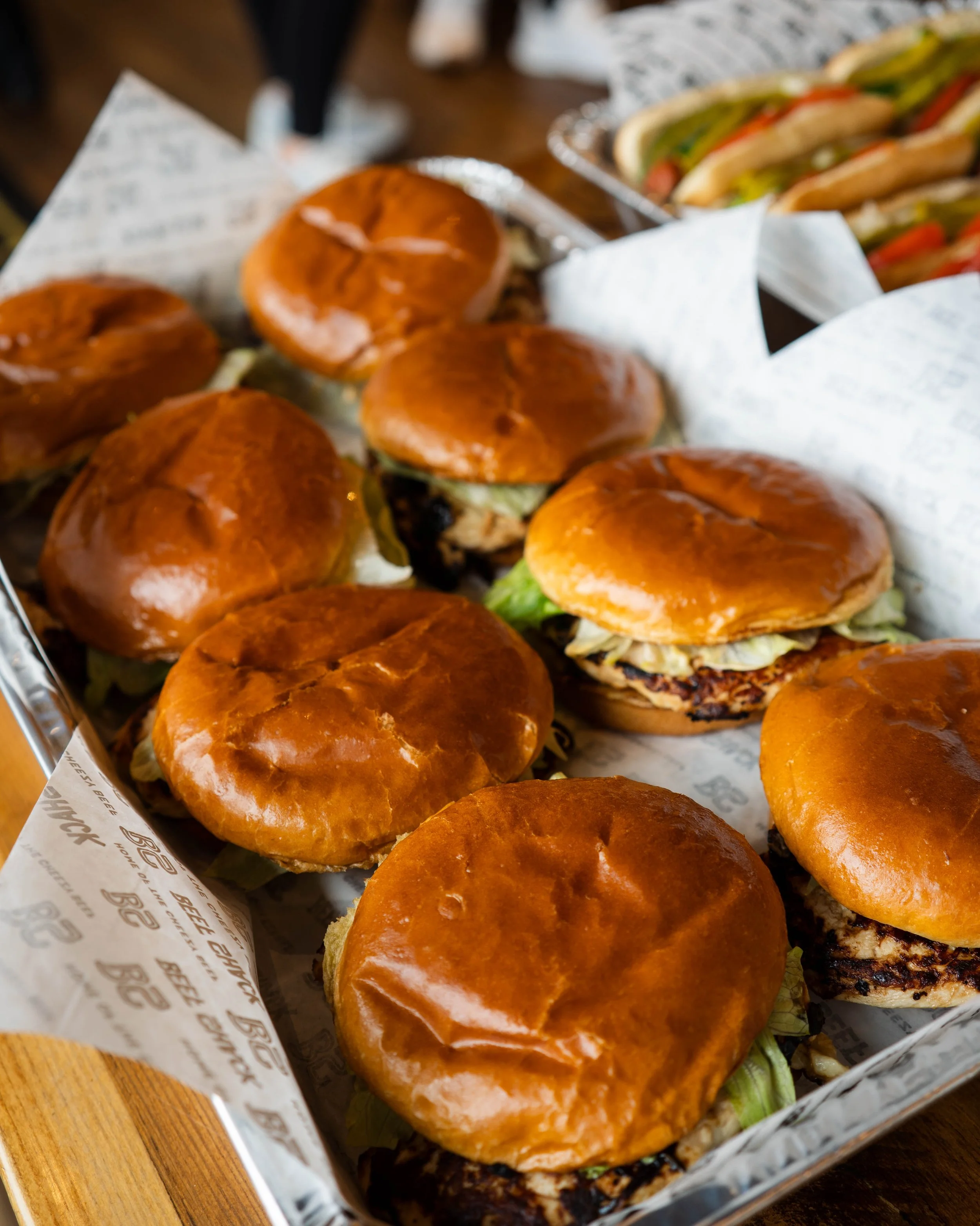 A tray of freshly made mini cheeseburgers with shiny brioche buns, lettuce, and grilled patties, with a side of sandwich-packed hot dogs in the background.