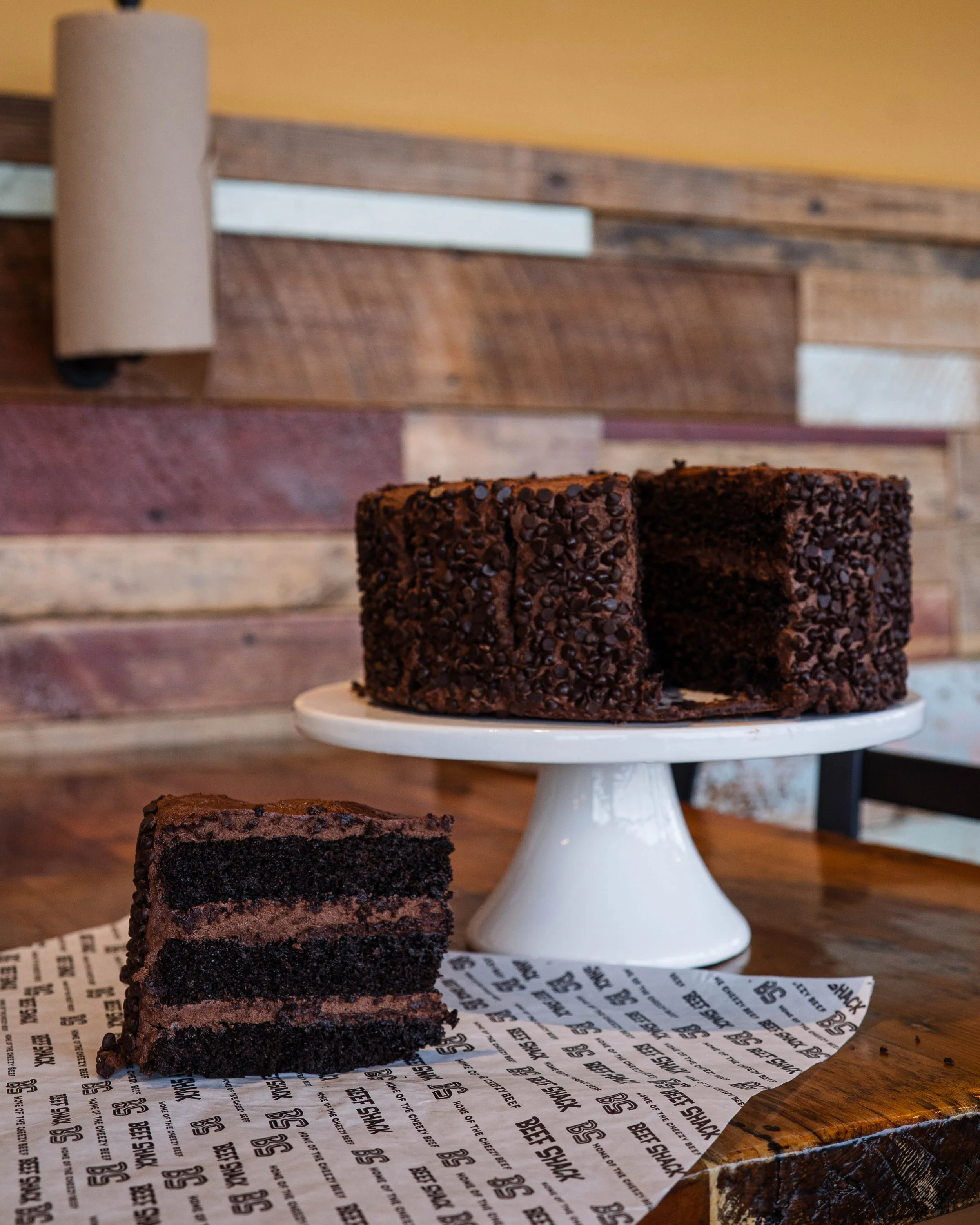 Chocolate cake with chocolate sprinkles on top, with a slice cut out, placed on a white cake stand in a rustic wooden setting.